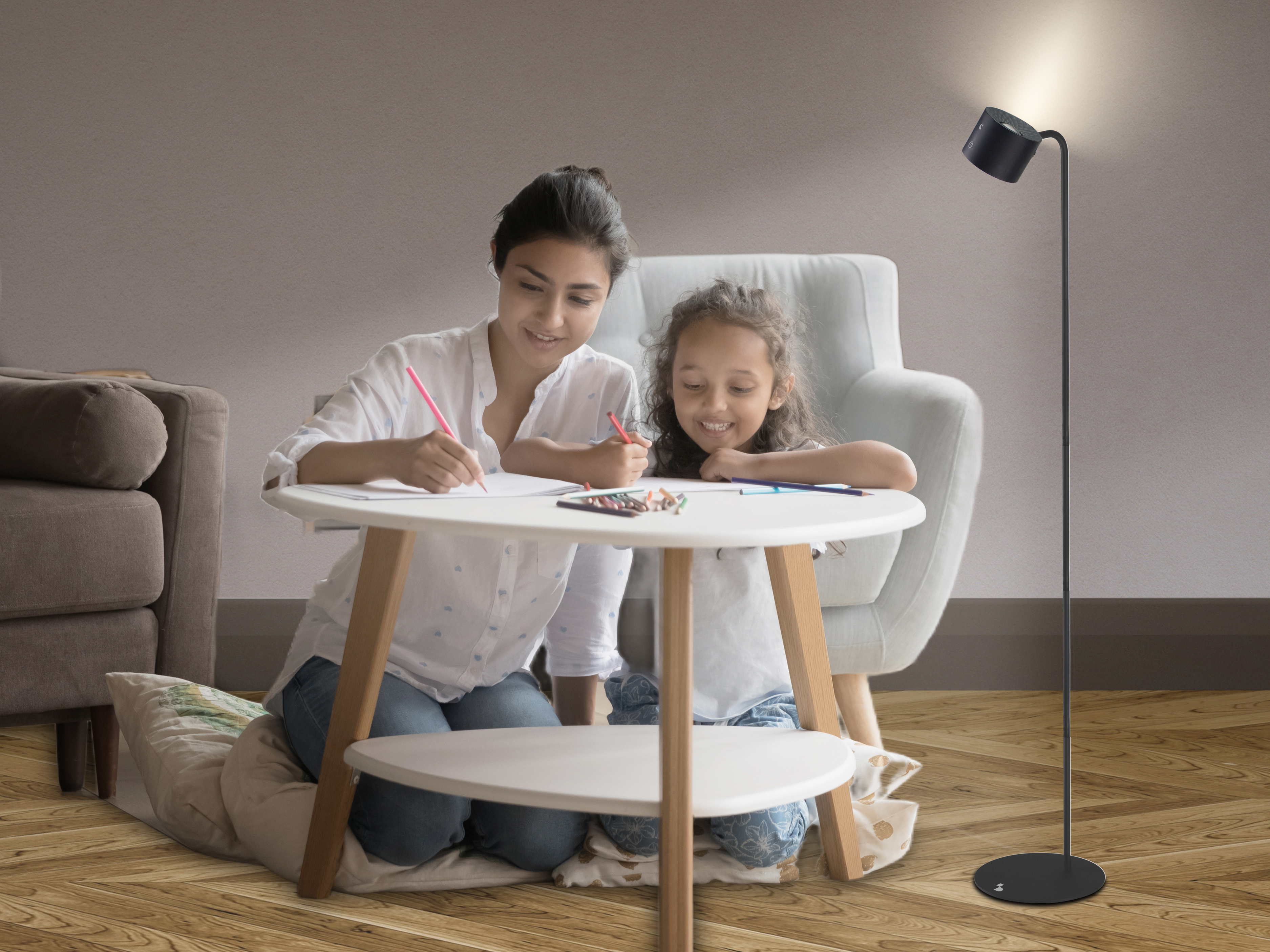 Una donna e una bambina disegnano a un tavolino. Una lampada e una poltrona sullo sfondo. Pavimento in legno.