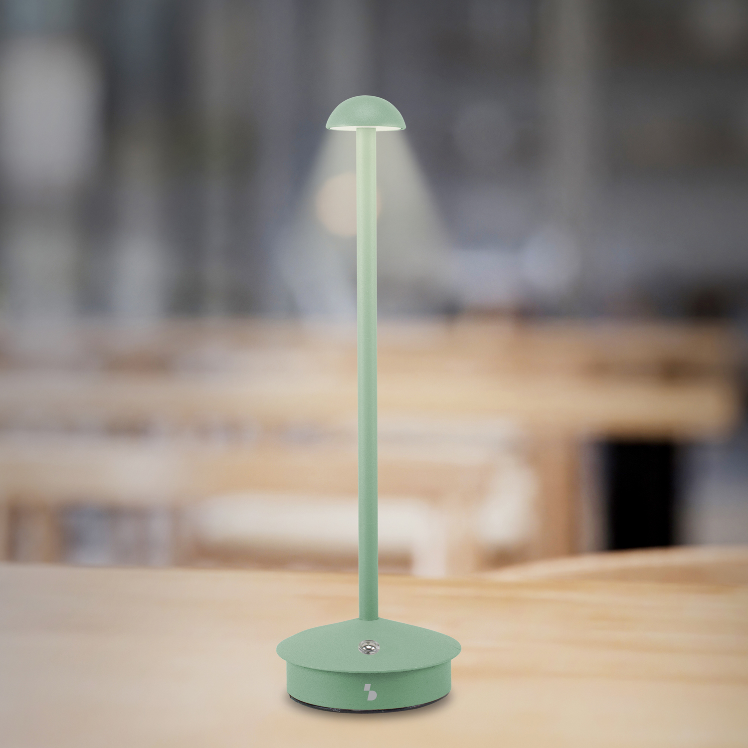 Una lampada da tavolo verde menta con parte superiore arrotondata e stelo verticale, posizionata su un tavolo di legno. Sfondo sfocato con mobili in legno.