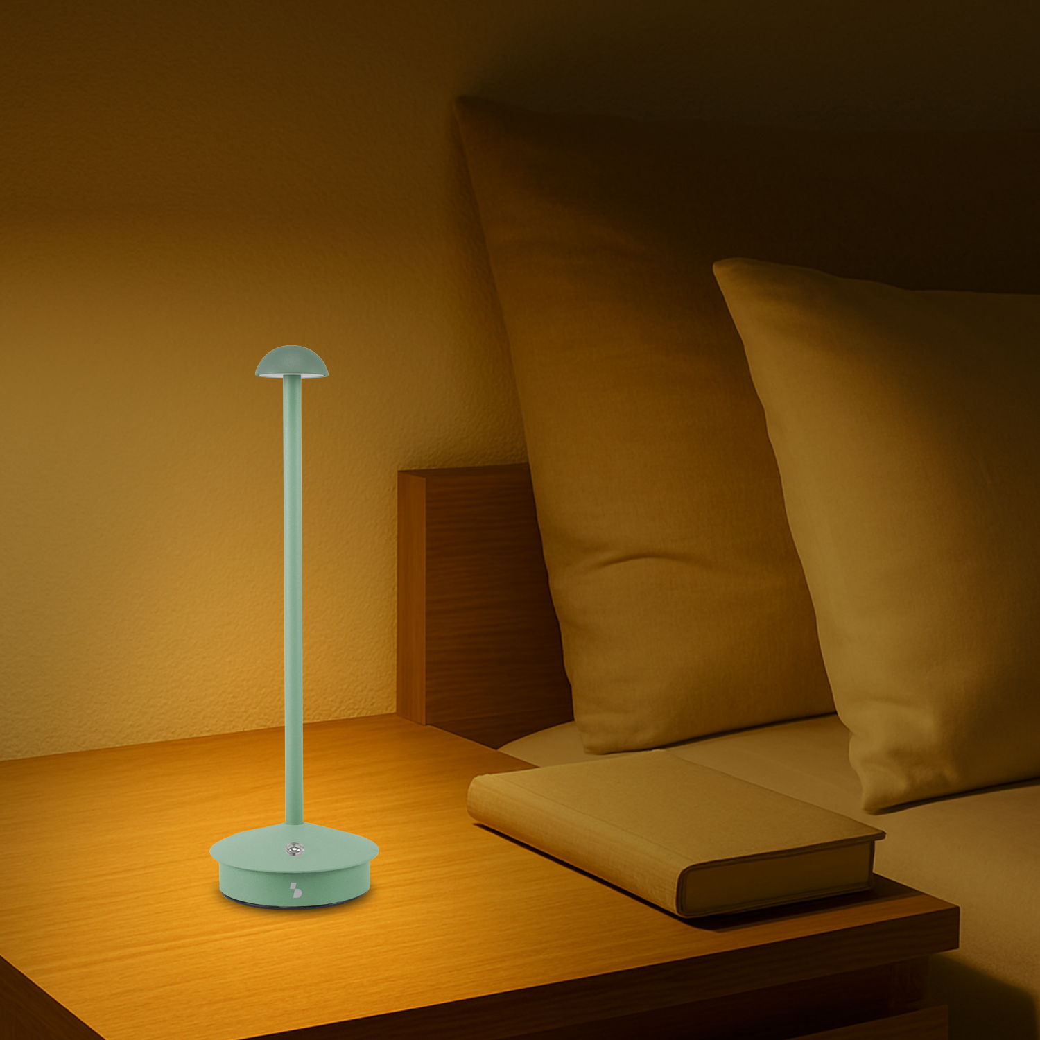 Una lampada verde menta su un comodino in legno. Un libro accanto. Cuscini sullo sfondo.