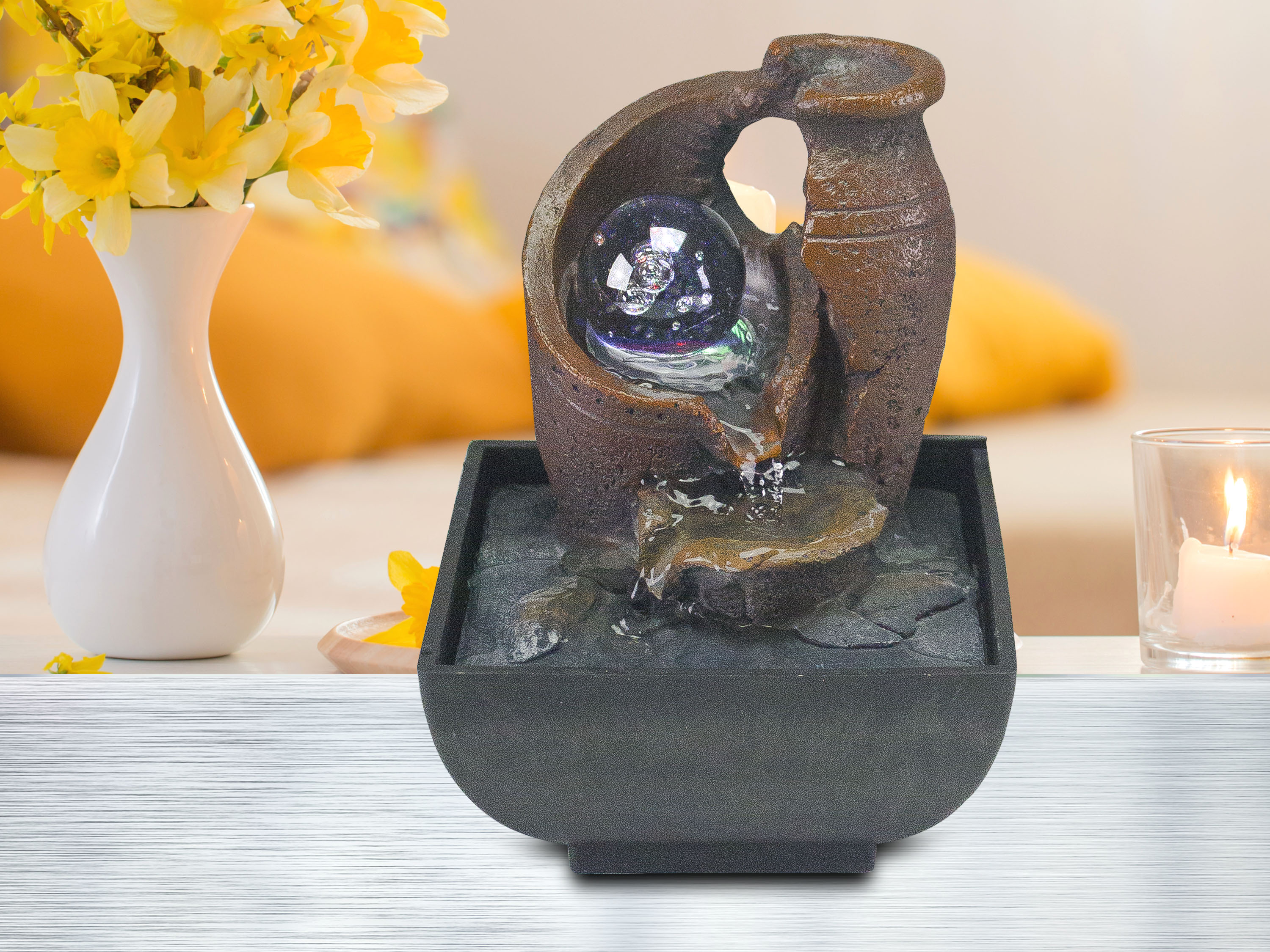 Fontana decorativa con sfera di vetro, fiori gialli in vaso, candela a destra.