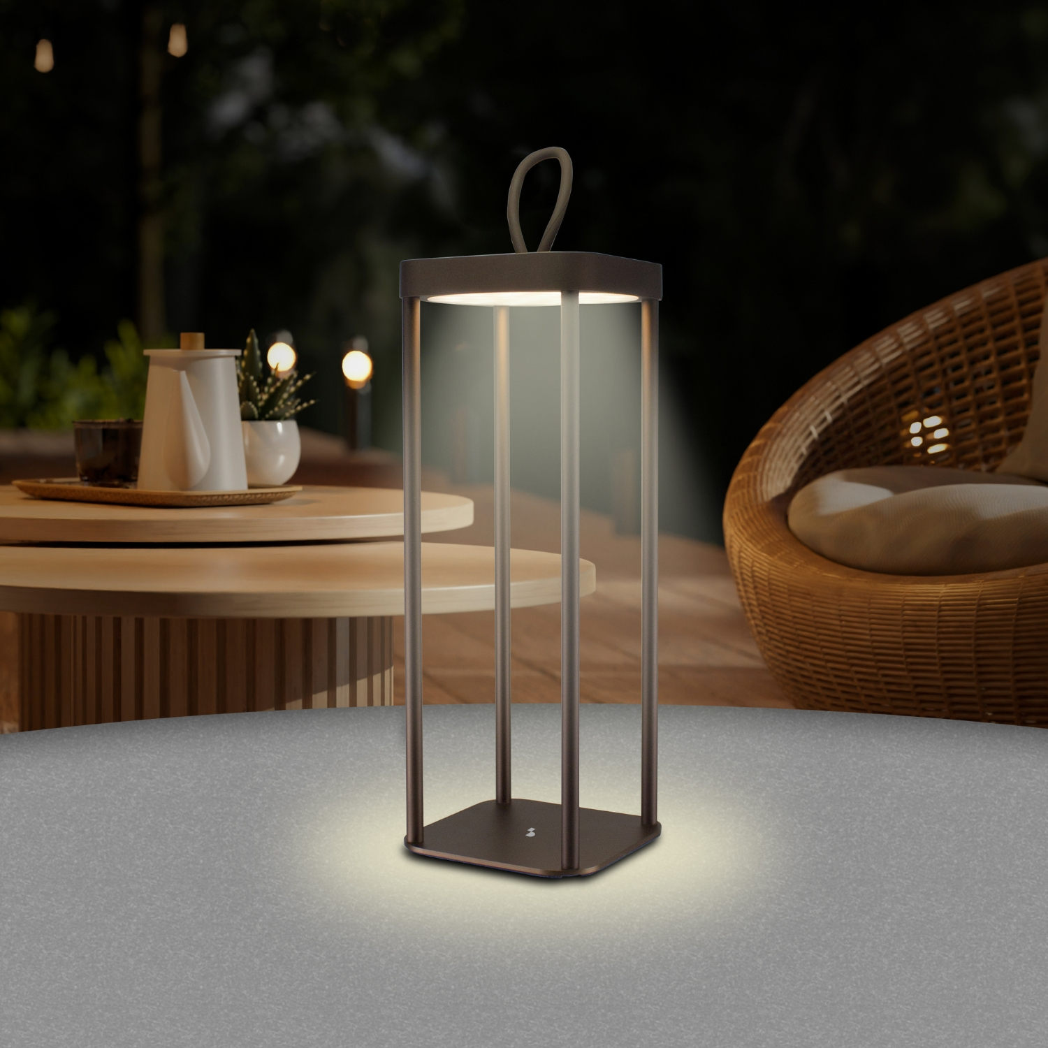 Una lampada quadrata illuminata con manico è su un tavolo. Una sedia in vimini e un tavolo con piante sono sullo sfondo.