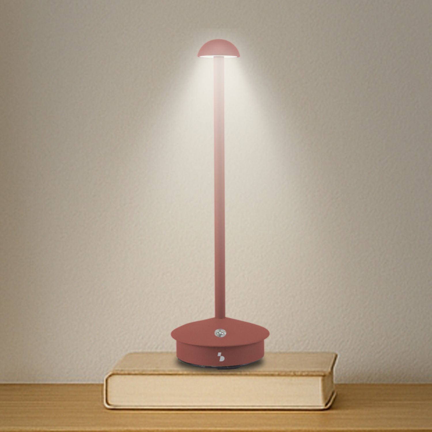 Una lampada da scrivania rossa è su un libro. La lampada è accesa, proietta un fascio di luce. Lo sfondo è una parete beige.