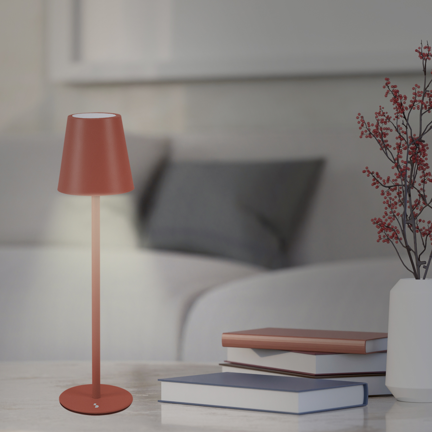 Una lampada in terracotta è su un tavolo vicino a libri e un vaso con bacche rosse. Un divano sullo sfondo sfocato.