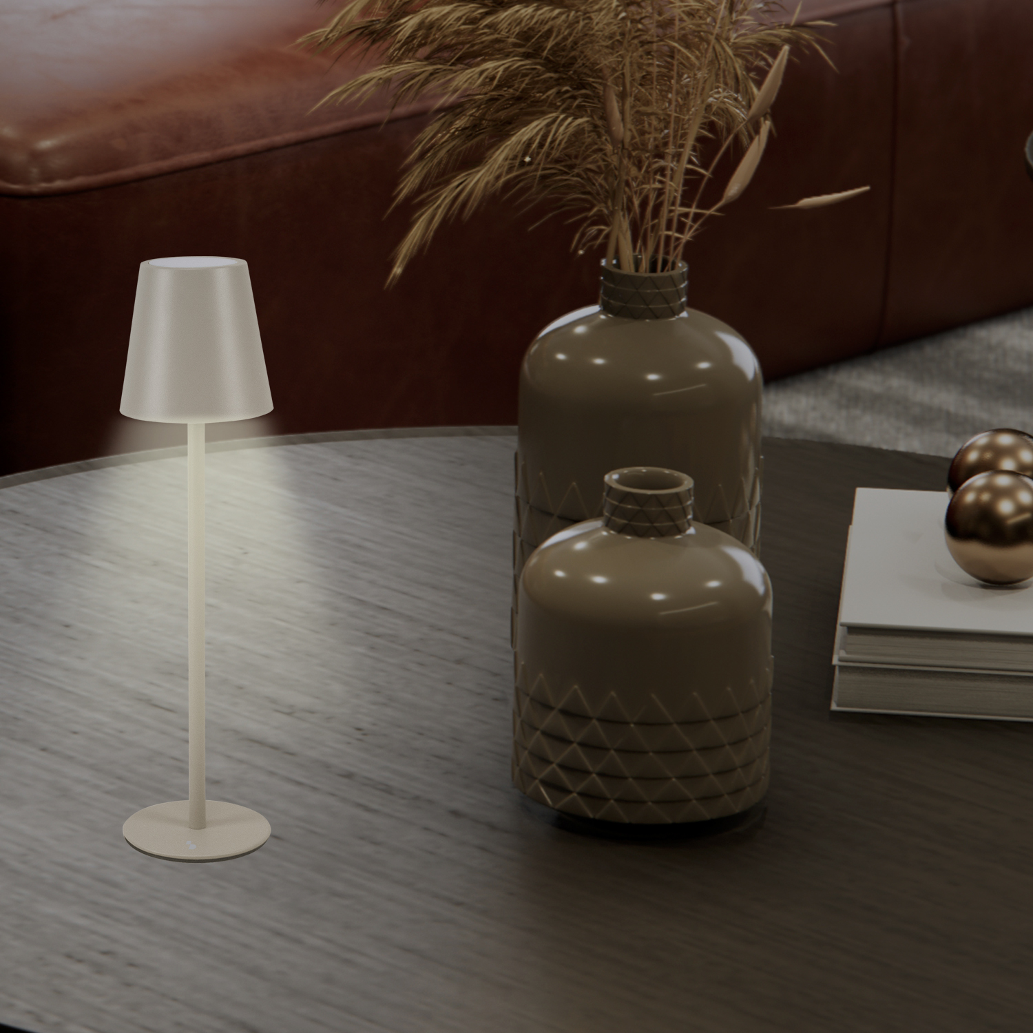 Una lampada da tavolo, vasi con piante essiccate e libri su un tavolo di legno. Un divano in pelle sullo sfondo.