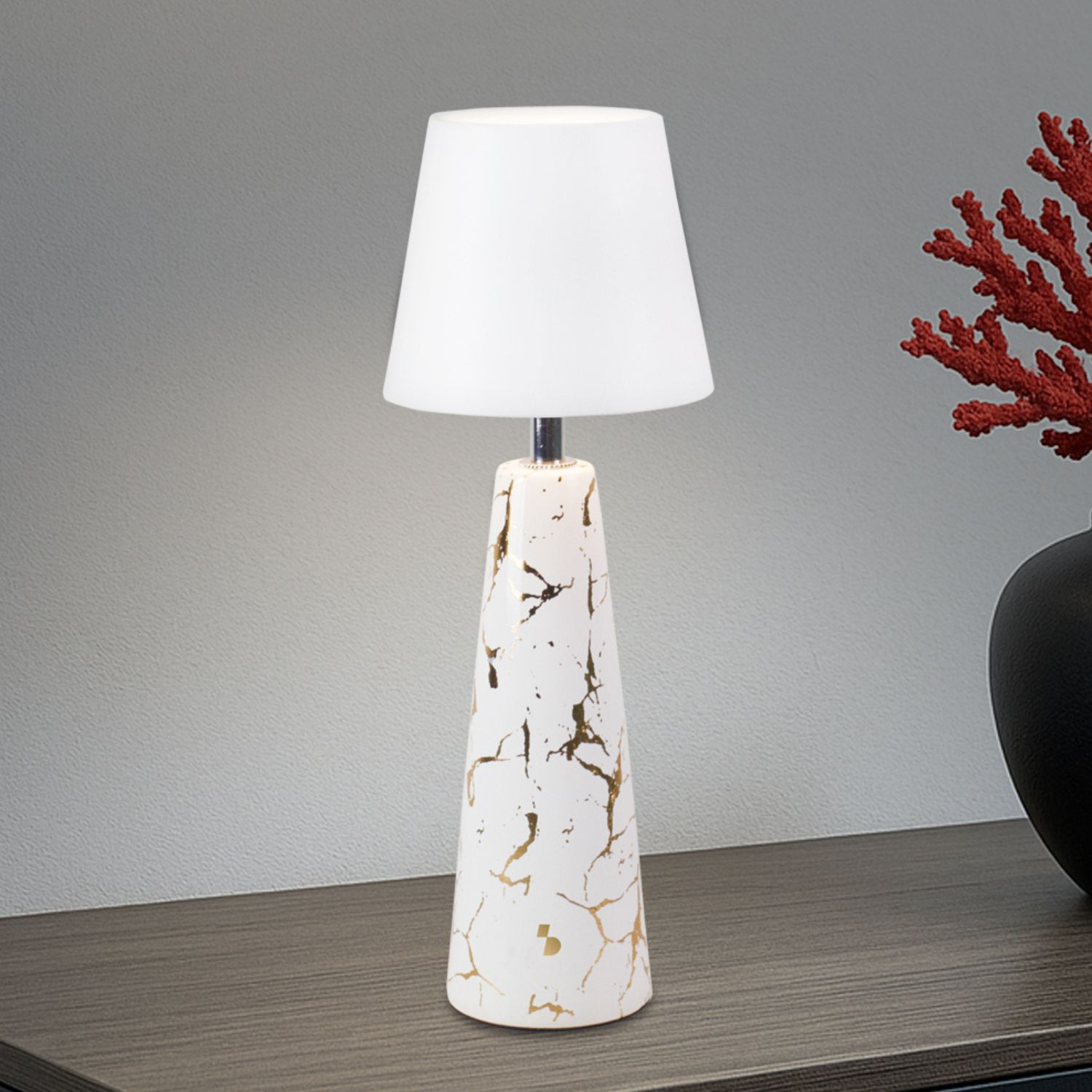 Una lampada bianca con base effetto marmo e paralume su una superficie in legno. Corallo rosso e vaso nero sullo sfondo.