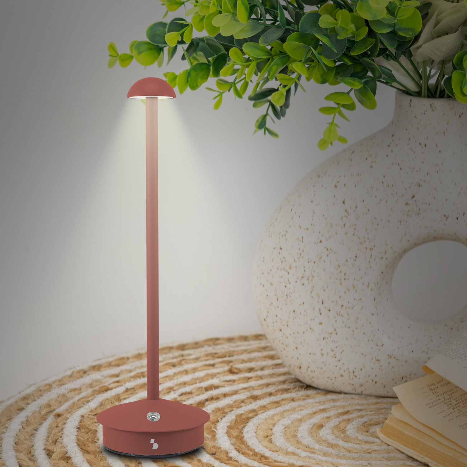 Una moderna lampada da scrivania con paralume rotondo. È rossa, con stelo beige, su un tappetino beige. Un vaso bianco con foglie verdi sullo sfondo.