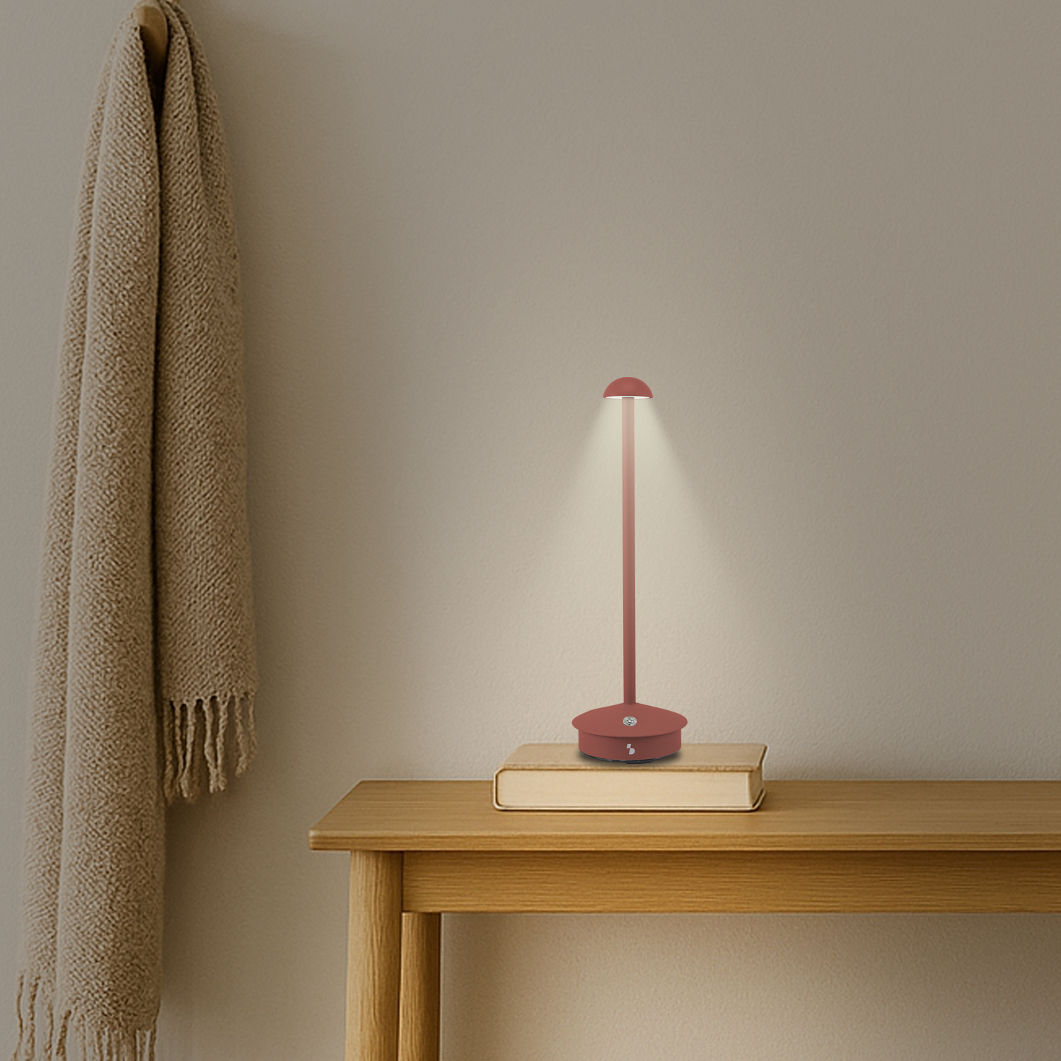Una lampada da scrivania rossa su un tavolo di legno. Un libro sotto. A sinistra una coperta beige. Lo sfondo è una parete beige.