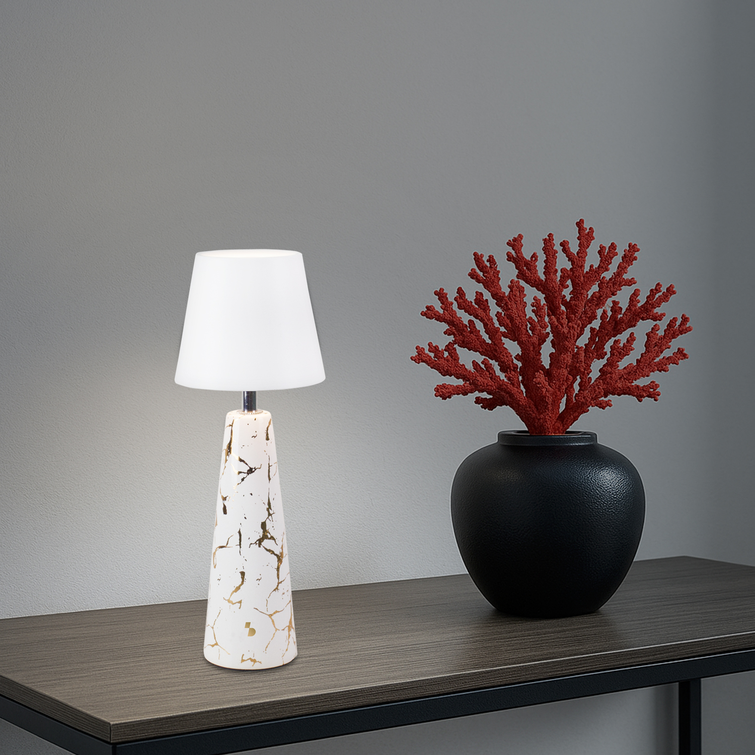Una lampada da tavolo con paralume bianco e base bianca con venature dorate, accanto a un vaso nero con corallo rosso.