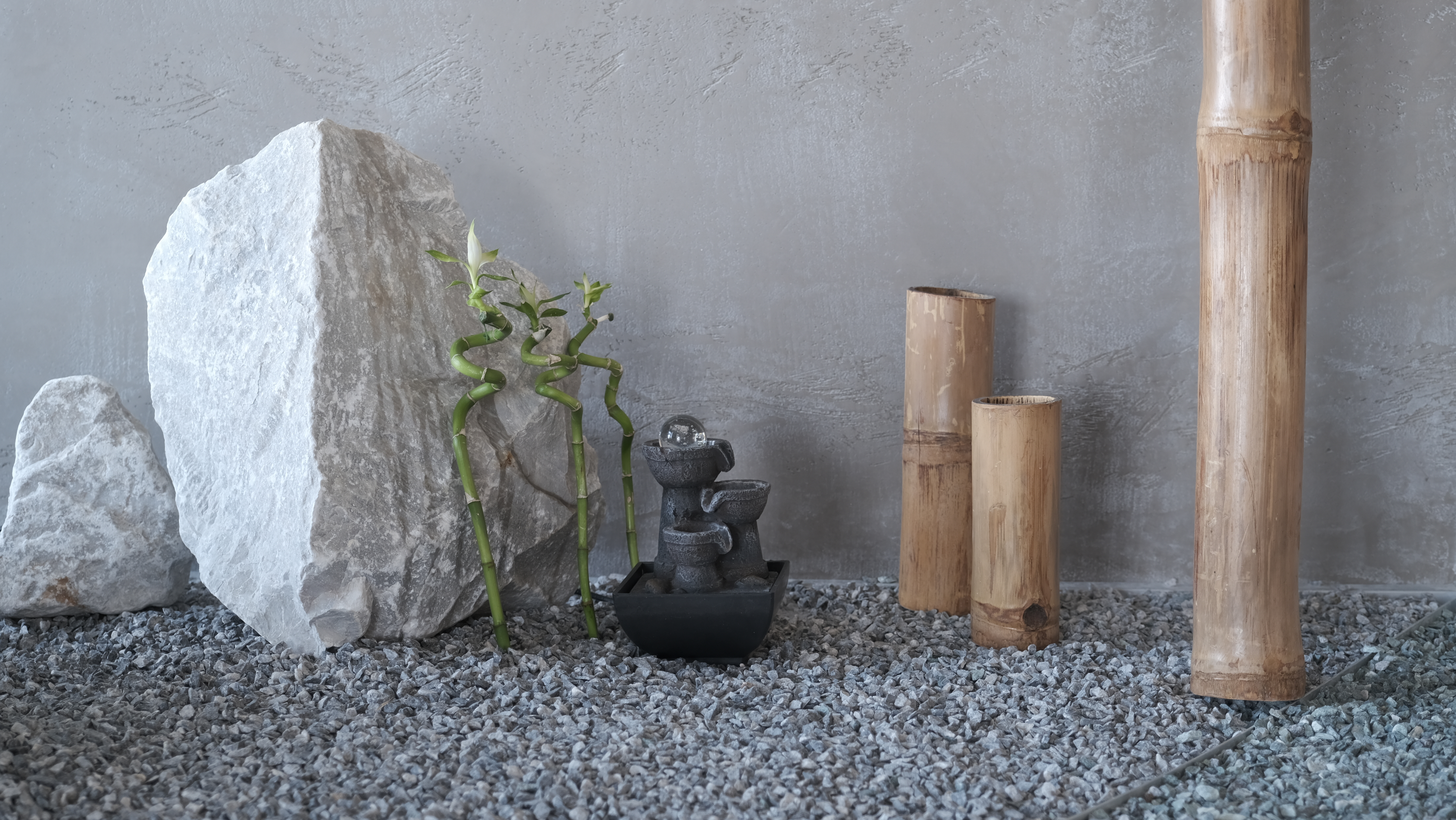 Una scena di giardino zen con rocce, bambù e una piccola fontana su ghiaia. Lo sfondo è un muro grigio strutturato.