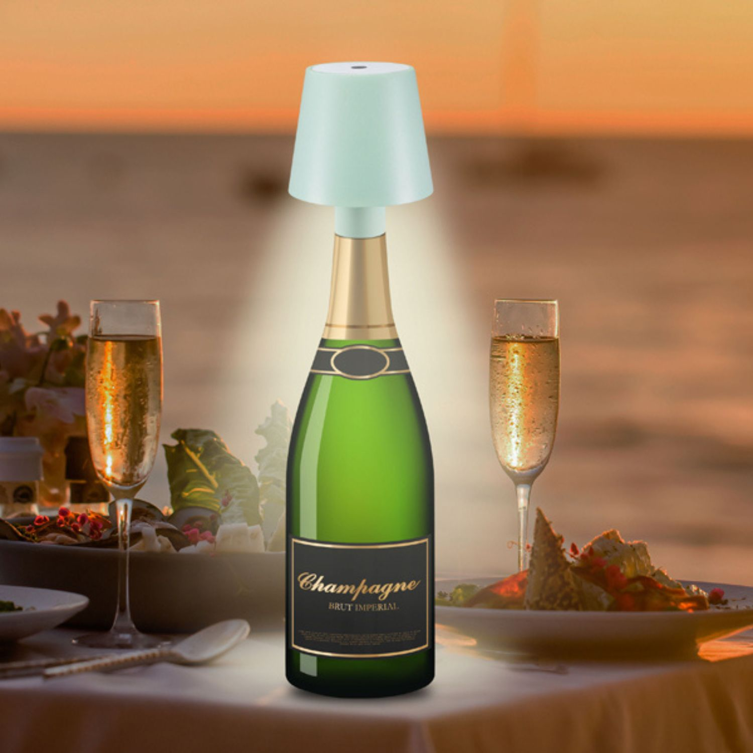 Una bottiglia di champagne con una lampada sopra, due bicchieri di champagne e cibo su un tavolo, sfondo tramonto.