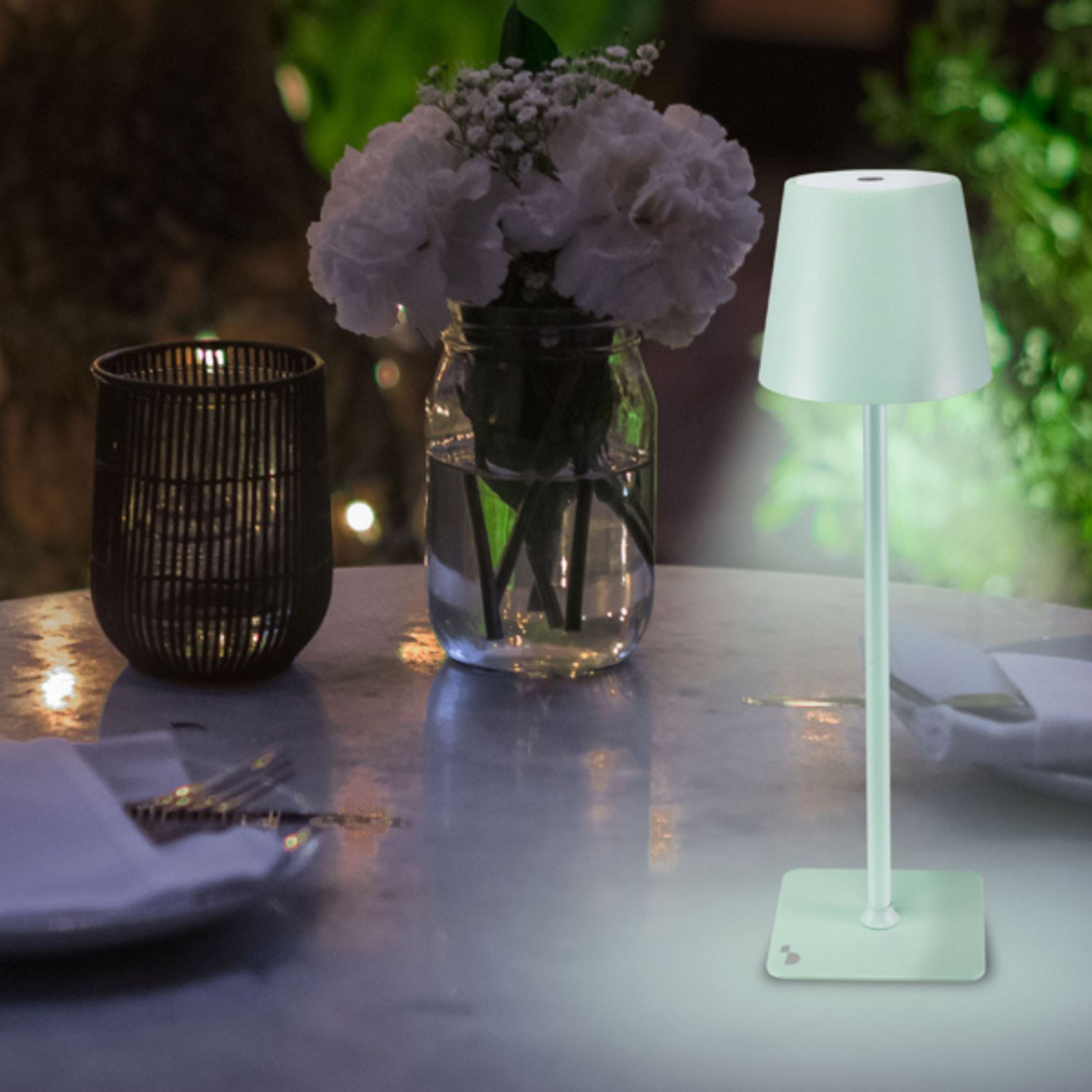 Tavolo con lampada, fiori e portacandele. La lampada è accesa. Scena serale con sfondo sfocato.