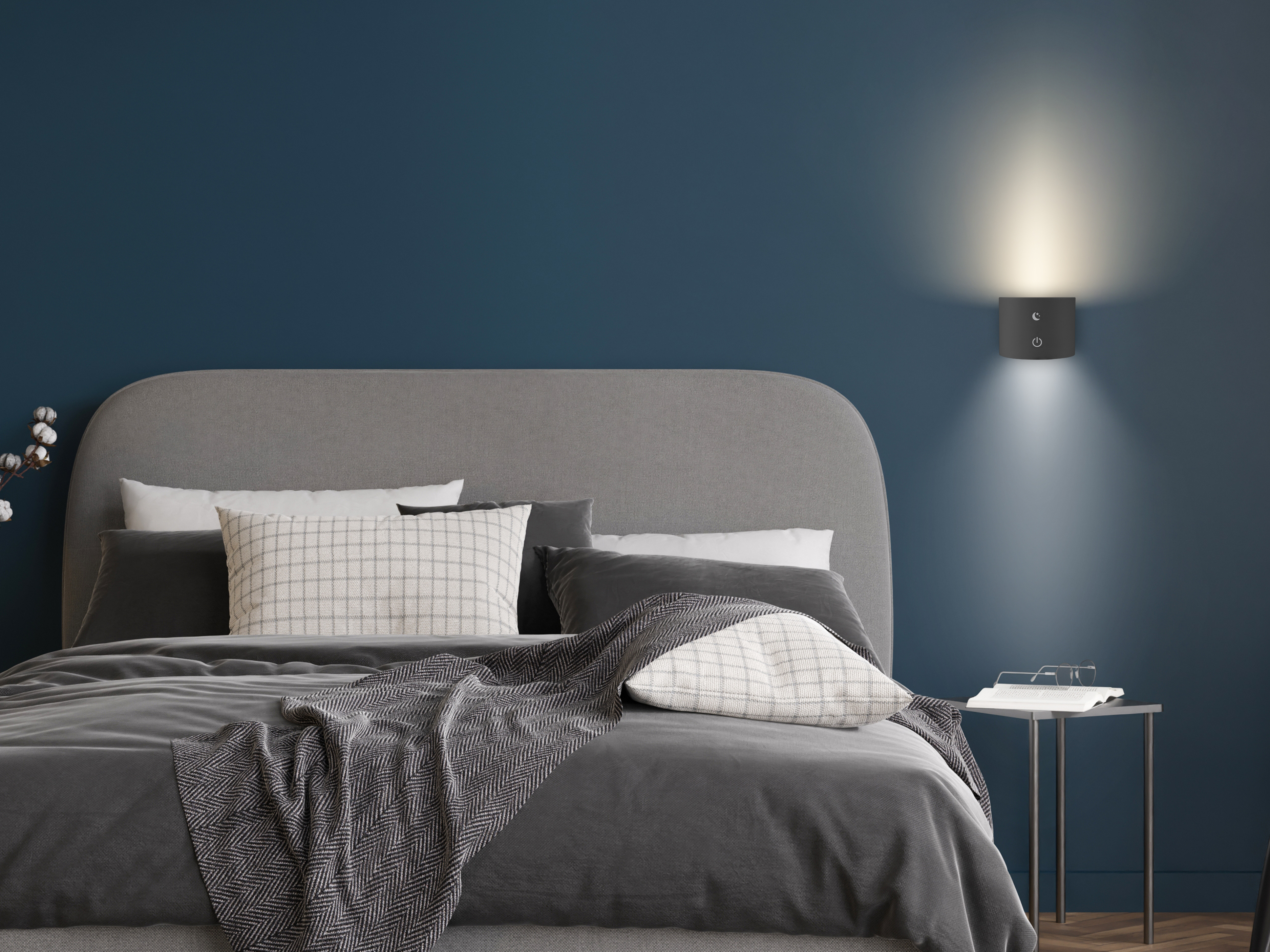 Una camera da letto con letto, cuscini e coperta. Una lampada da parete è accesa, illuminando lo spazio. Tavolino.