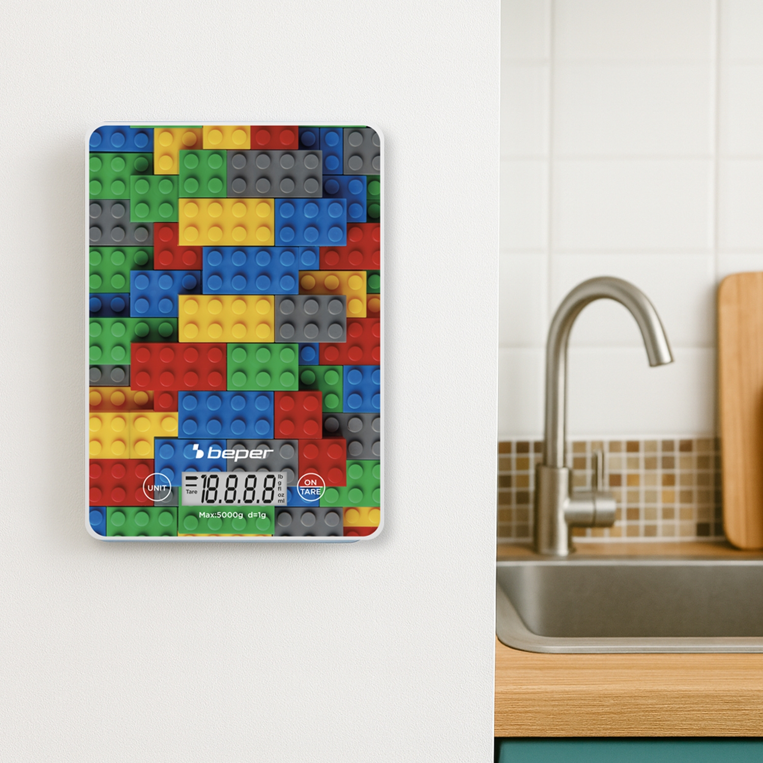 Una bilancia da cucina con design a mattoncini Lego è appesa a una parete bianca. Sullo sfondo si vedono un rubinetto, un lavandino e un tagliere.