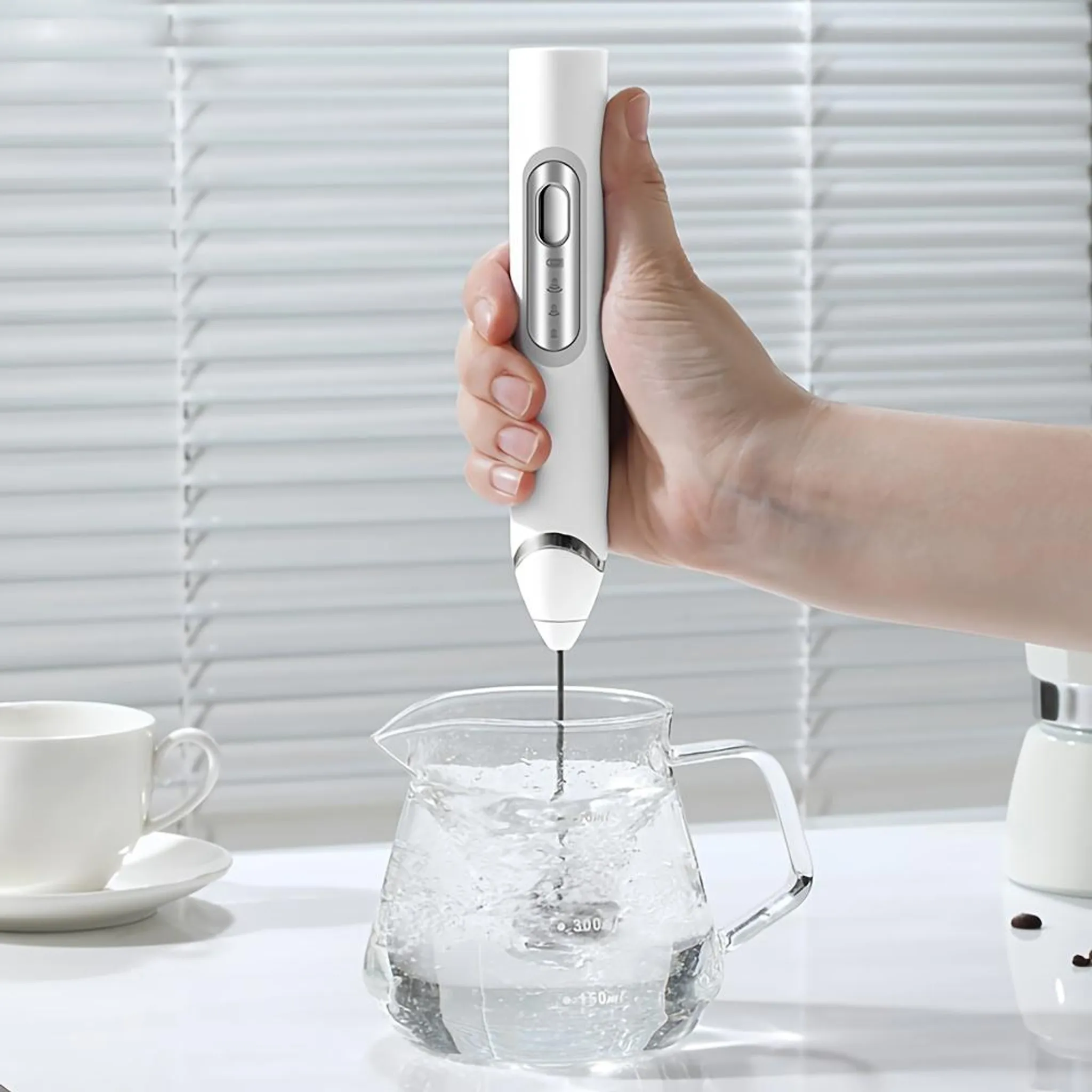 Eine Hand hält einen weißen Milchaufschäumer über einem Glaskrug mit Wasser. Im Hintergrund befinden sich Jalousien. Auf dem Tisch steht eine Tasse.