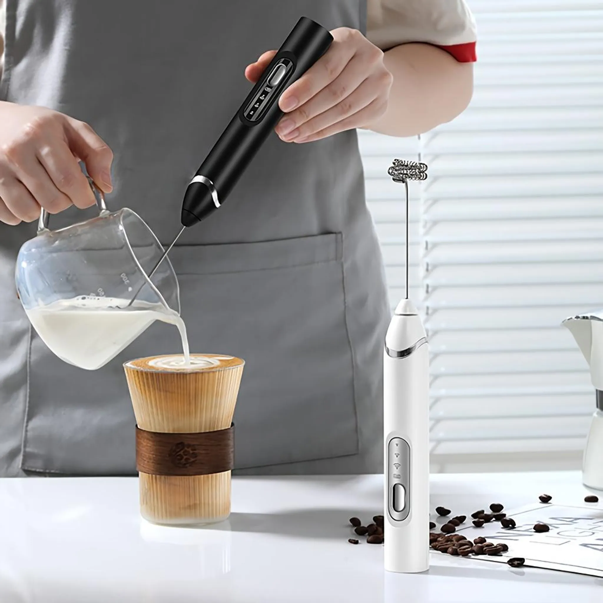 Eine Person gießt Milch in eine Kaffeetasse. Zwei Aufschäumer sind sichtbar. Kaffeebohnen liegen auf dem Tisch.
