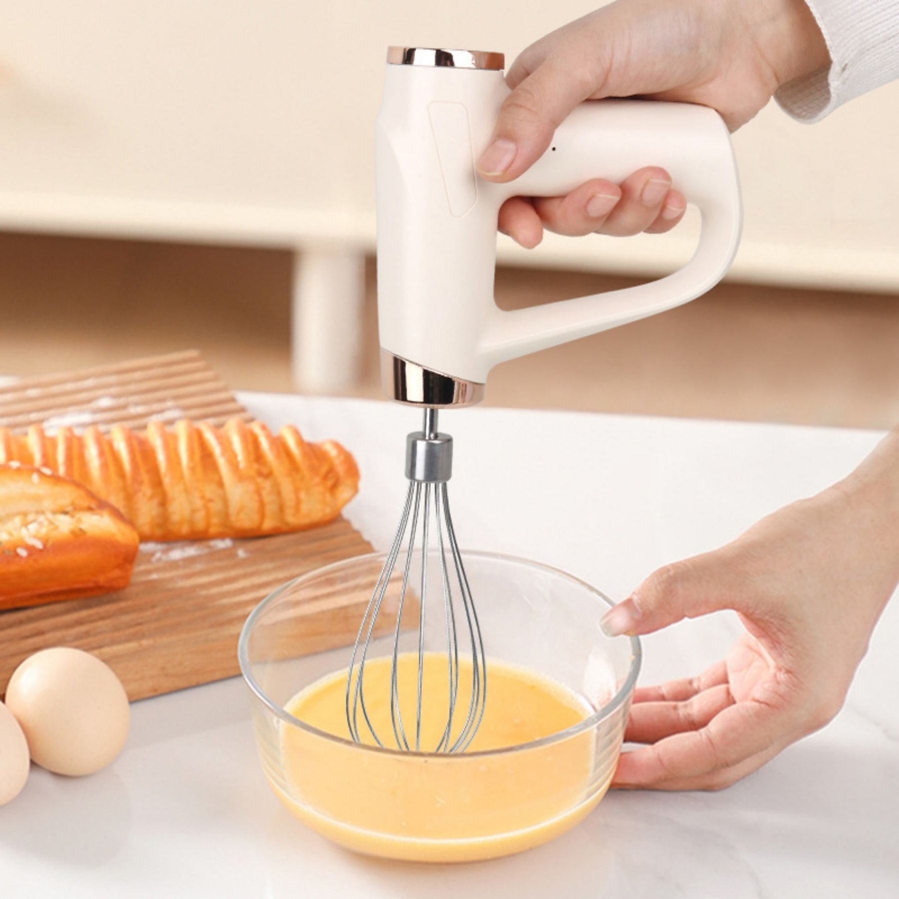 Eine Hand hält einen weißen Mixer über einer Schüssel mit gelber Flüssigkeit. Brot und Eier liegen auf dem Tisch.