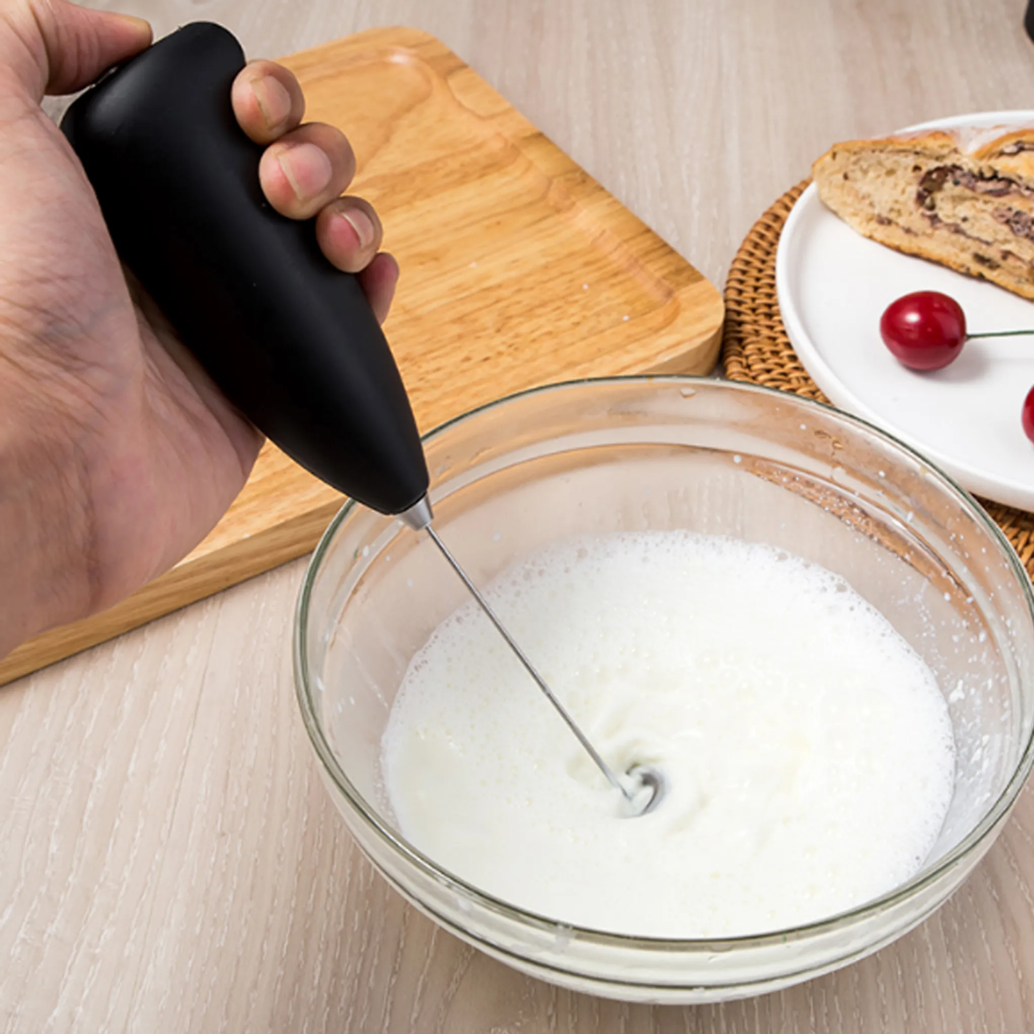 Hand hält einen schwarzen Milchaufschäumer über einer Schüssel Milch. Ein Brotstück und Kirschen liegen auf einem Teller.