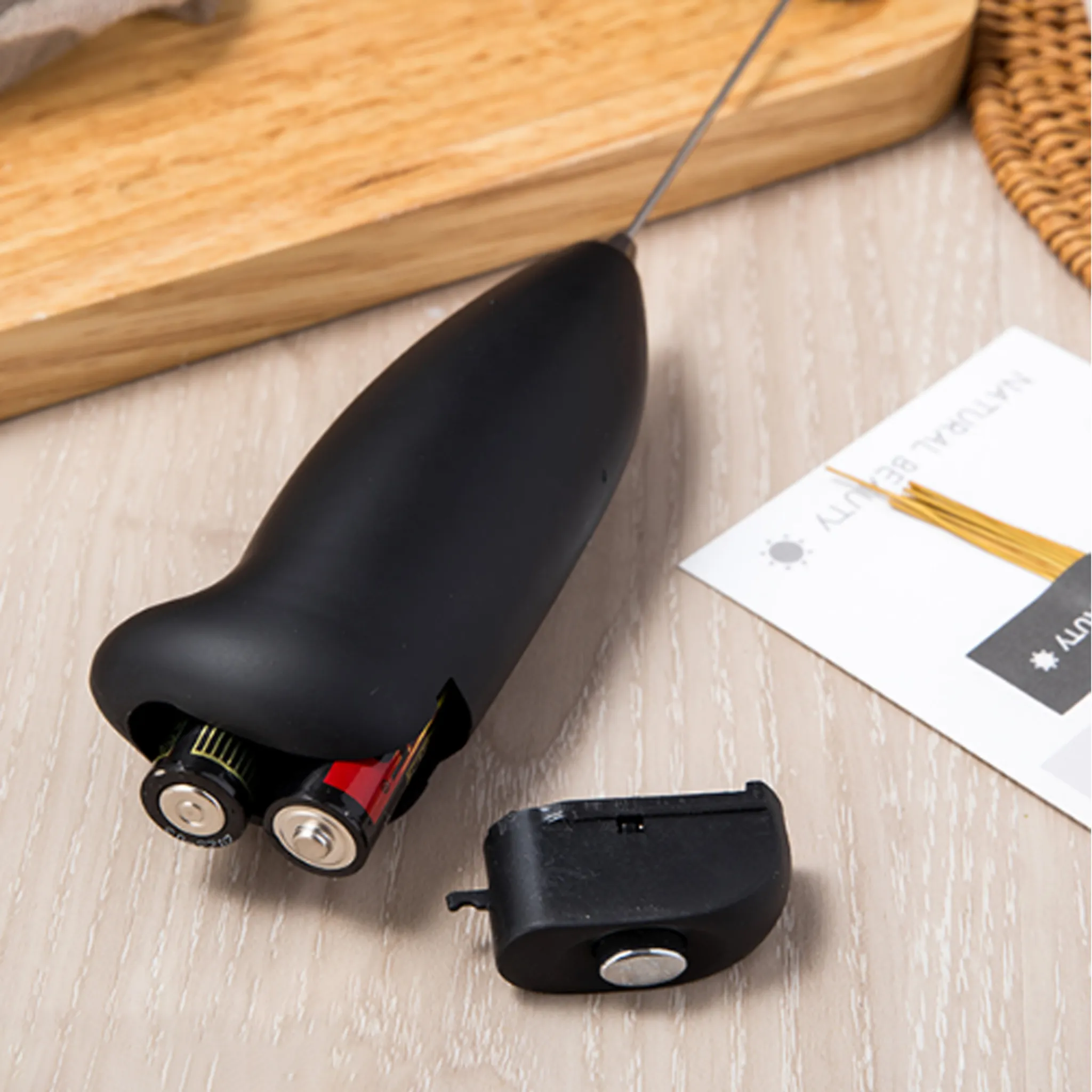 Ein schwarzer Milchaufschäumer mit Batterien, einem abgenommenen Deckel und einem Holzbrett im Hintergrund.