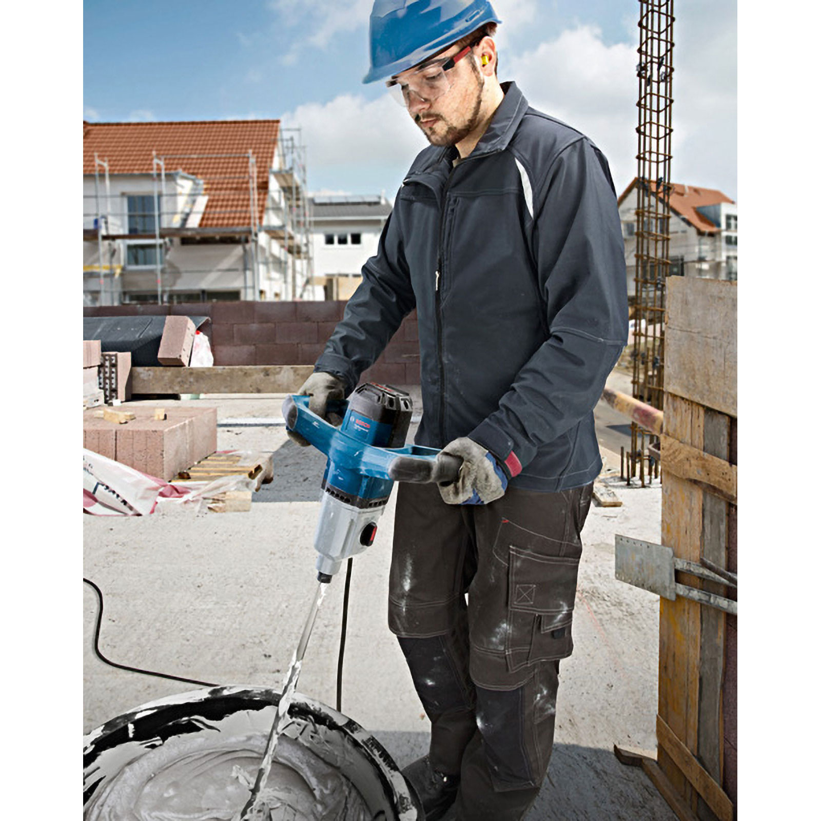 Un operaio edile utilizza un miscelatore per mescolare il materiale in un secchio. Ci sono case sullo sfondo. L'operaio indossa un casco blu.