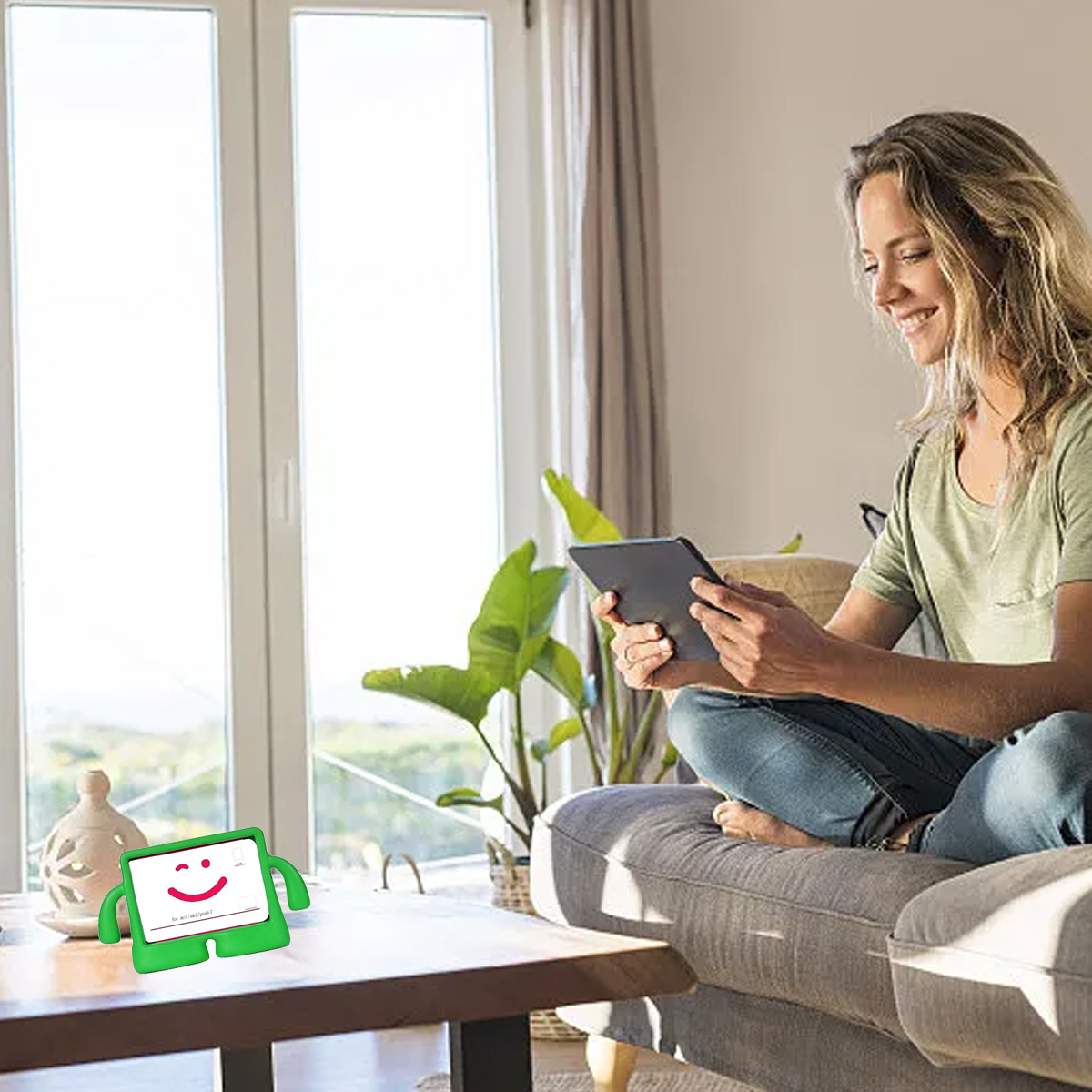 Eine Frau sitzt auf einem Sofa und benutzt ein Tablet. Eine grüne Cartoon-Figur steht auf einem Tisch. Fenster und Pflanzen sind im Hintergrund.
