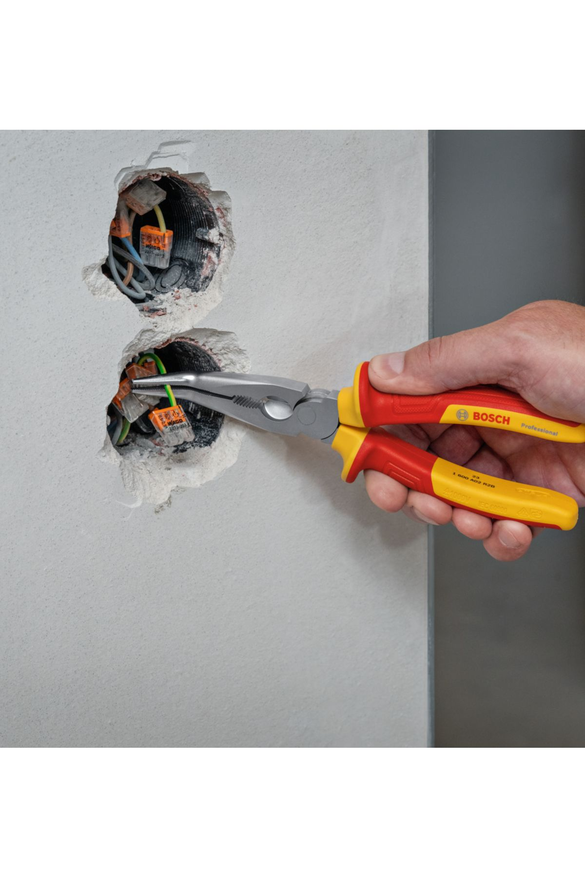 El, kablolarla duvar açıklıklarının yakınında Bosch pense tutuyor. Kırmızı ve sarı sap, gri pense. Elektrik işleri devam ediyor.