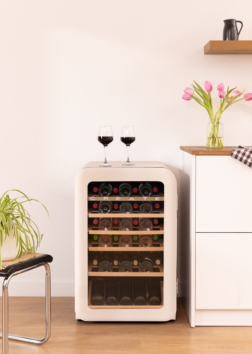 Una vinoteca con copas encima. Tulipanes en jarrón, botellas dentro. Planta y silla a la izquierda.