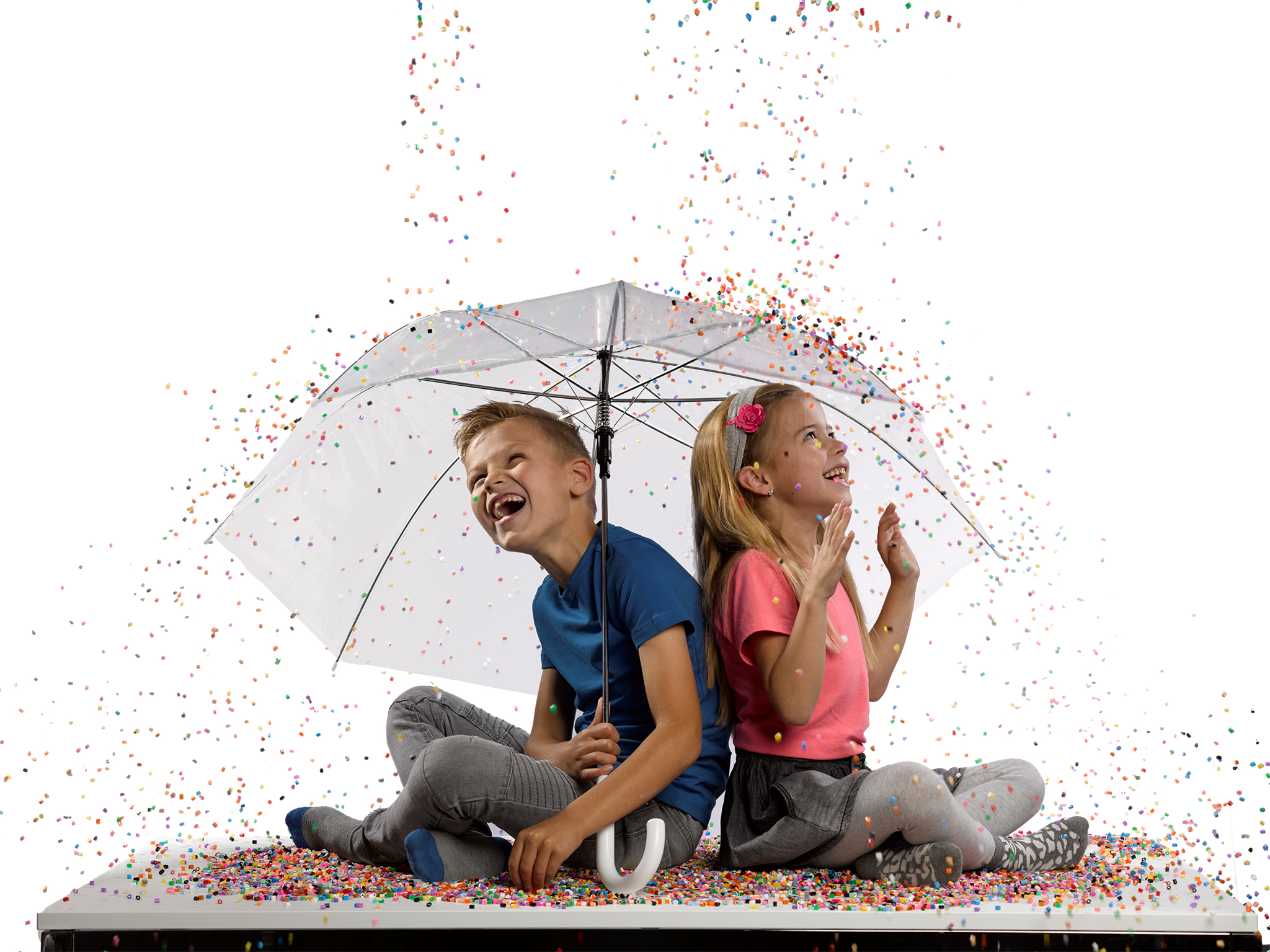 Zwei Kinder unter einem Regenschirm lachen, während Konfetti fällt. Sie sitzen auf einer Konfetti-bedeckten Oberfläche.