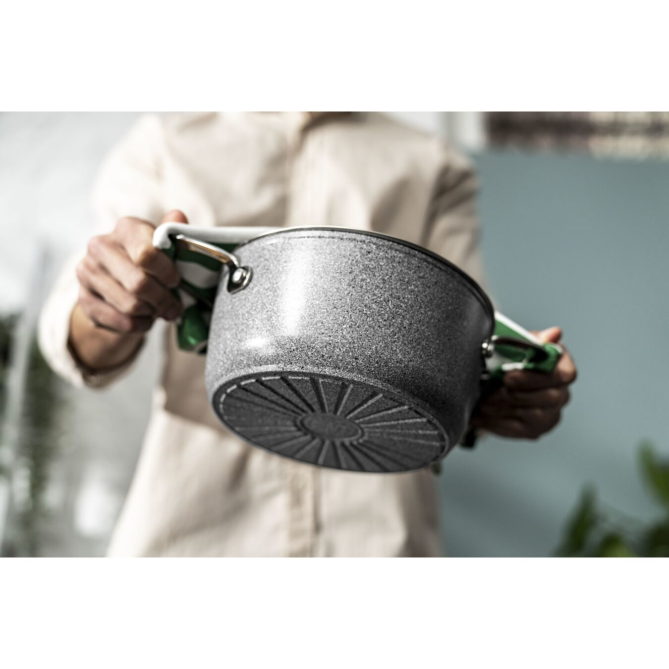 Una persona sostiene una olla gris moteada con asas. Paño a rayas verdes y blancas para sujetar la olla. Camisa beige en el fondo.