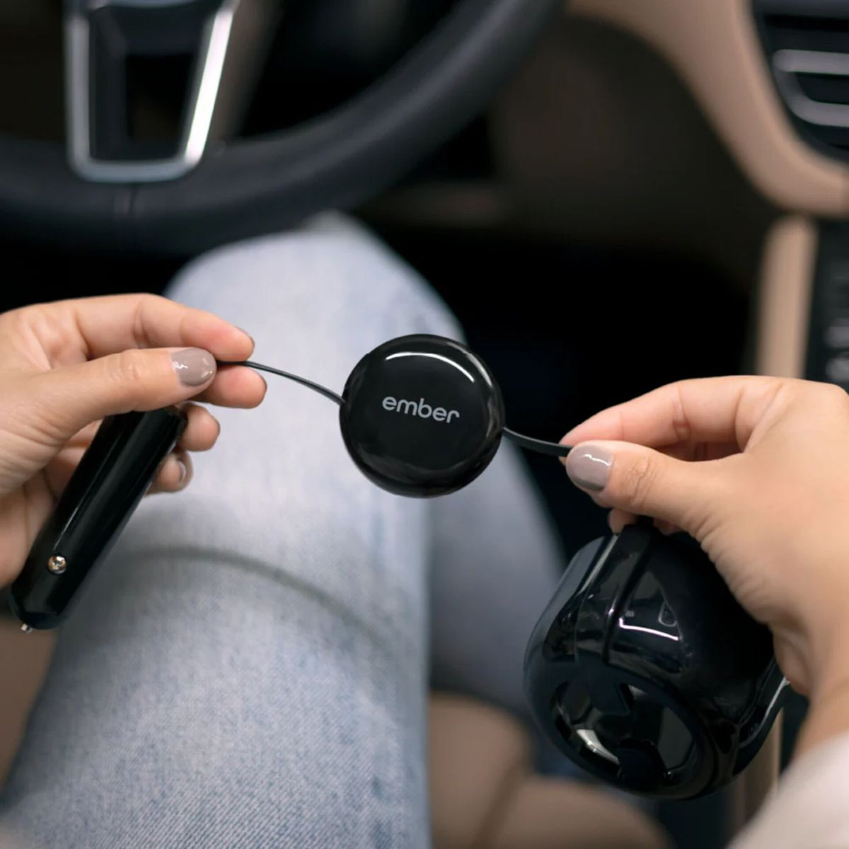 Hände halten ein schwarzes Ember-Gerät und einen Auto-Adapter in einem Auto. Lenkrad und Sitz sind im Hintergrund zu sehen.