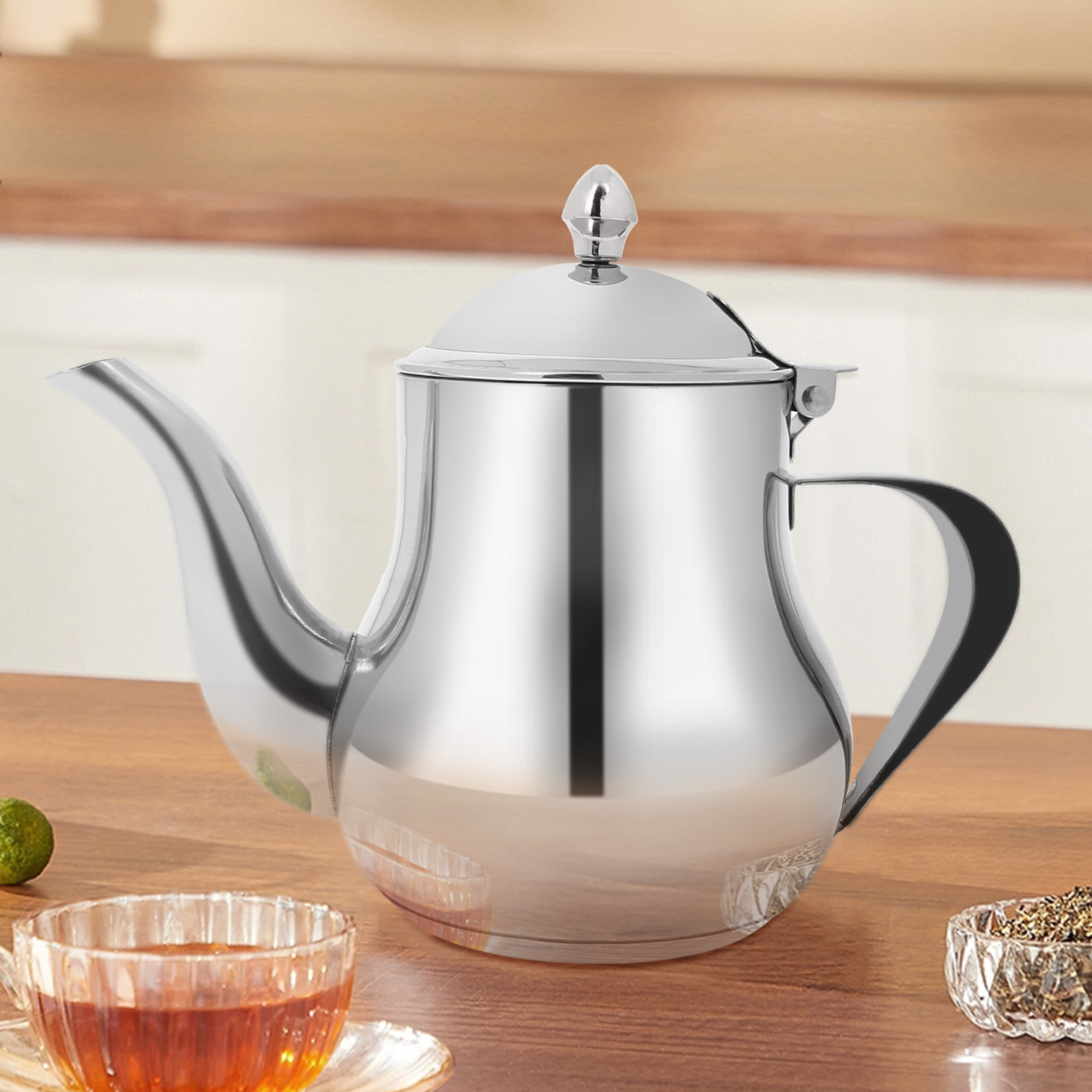 Eine glänzende silberne Teekanne mit Deckel steht auf einem Holztisch. Eine Tasse Tee und eine Schüssel mit Kräutern befinden sich im Vordergrund.
