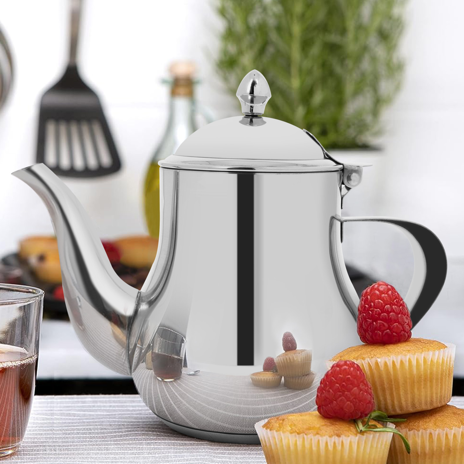Una tetera plateada brillante con frambuesas y pastelitos, en una mesa. Un vaso de té a la izquierda.