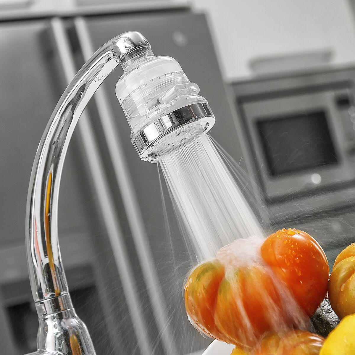 Ein Wasserhahn sprüht Wasser auf Tomaten in einer Küche. Die Tomaten sind rot und orange. Eine Mikrowelle und ein Kühlschrank sind im Hintergrund.