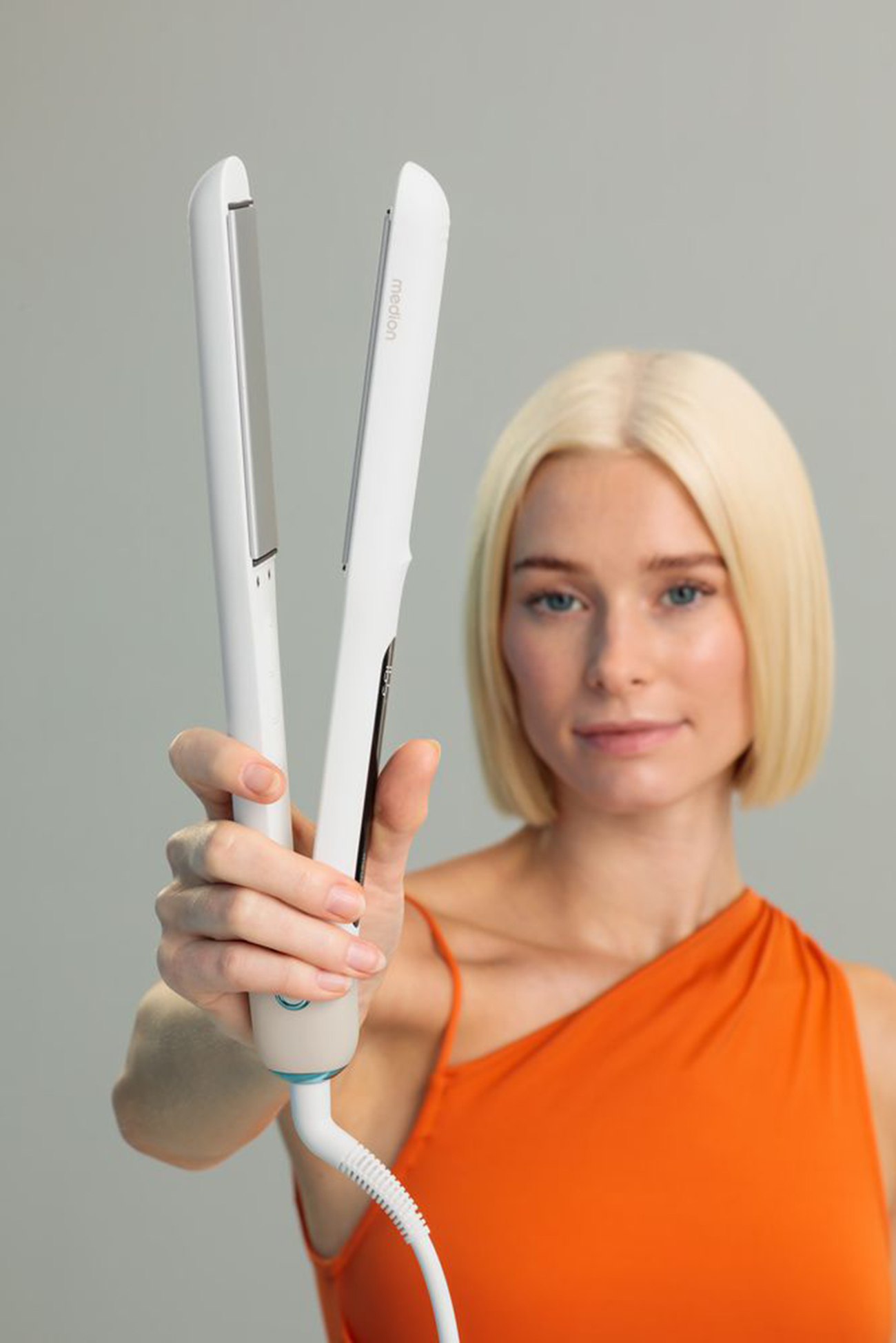 Blondynka trzyma białą prostownicę do włosów. Ma na sobie pomarańczowy top. Tło jest szare.