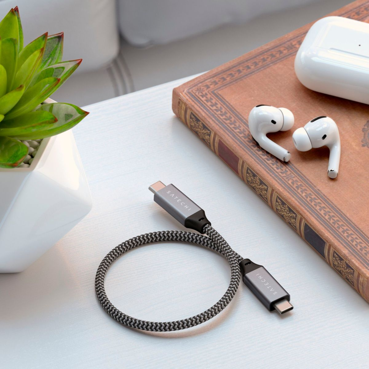 Pleciony kabel USB-C z szarymi złączami i czarno-szarym kablem. Widoczne są również słuchawki AirPods i sukulent.