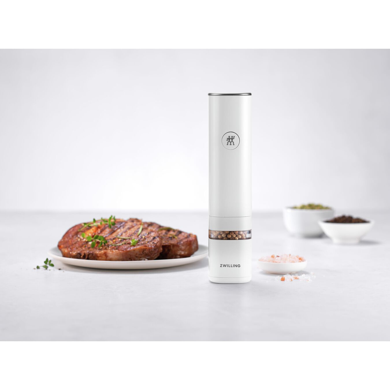 Un molinillo de pimienta eléctrico blanco, bistec y sal sobre una mesa. Cuencos con especias al fondo. La escena está bien iluminada.