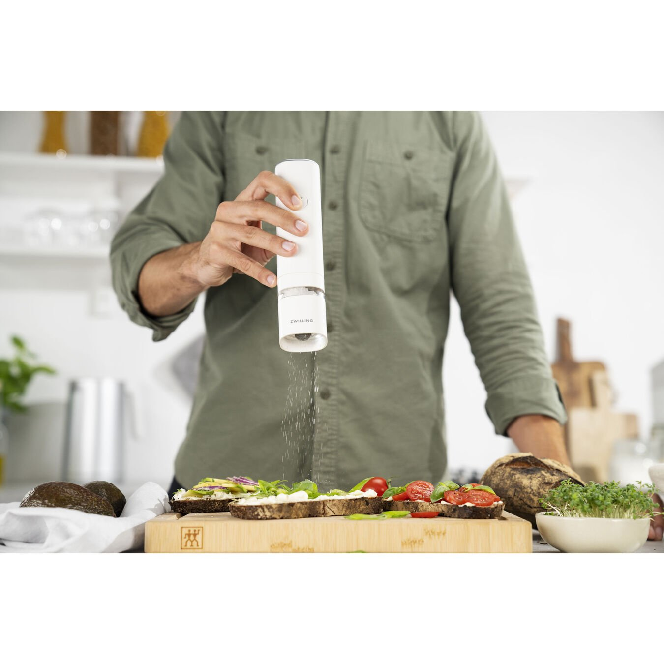 Una persona espolvorea condimento sobre un sándwich. Los ingredientes incluyen tomates, aguacate y brotes. Se usa un molinillo blanco.