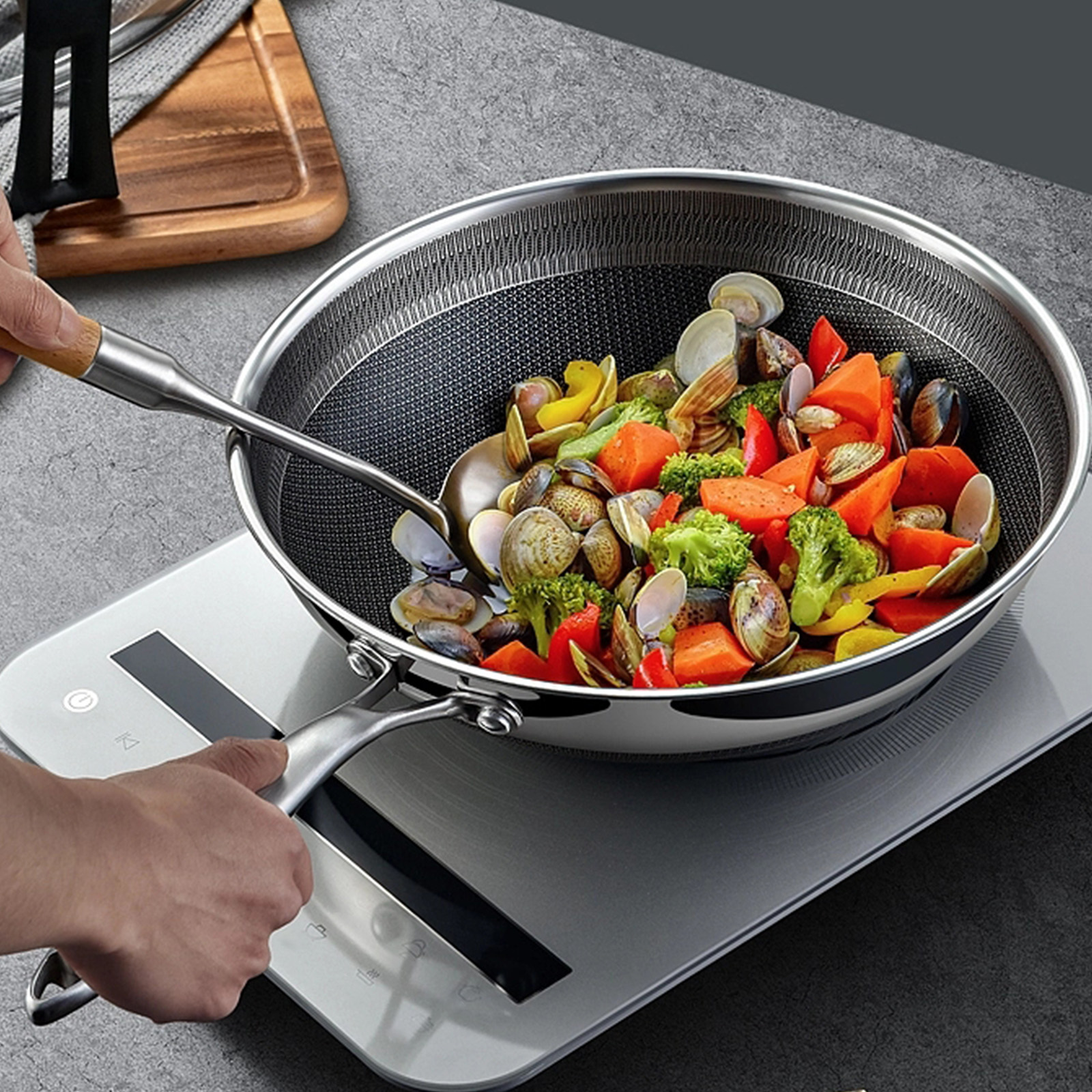 Un wok avec des légumes et des palourdes est sur une plaque à induction. Une main tient une cuillère et l'autre la poignée du wok.