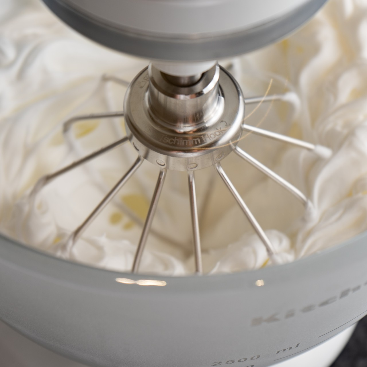Nahaufnahme eines Mixers, der weiße Sahne in einer Schüssel verrührt. Der Mixer ist silberfarben, die Schüssel hellgrau. Sahne steht im Fokus.