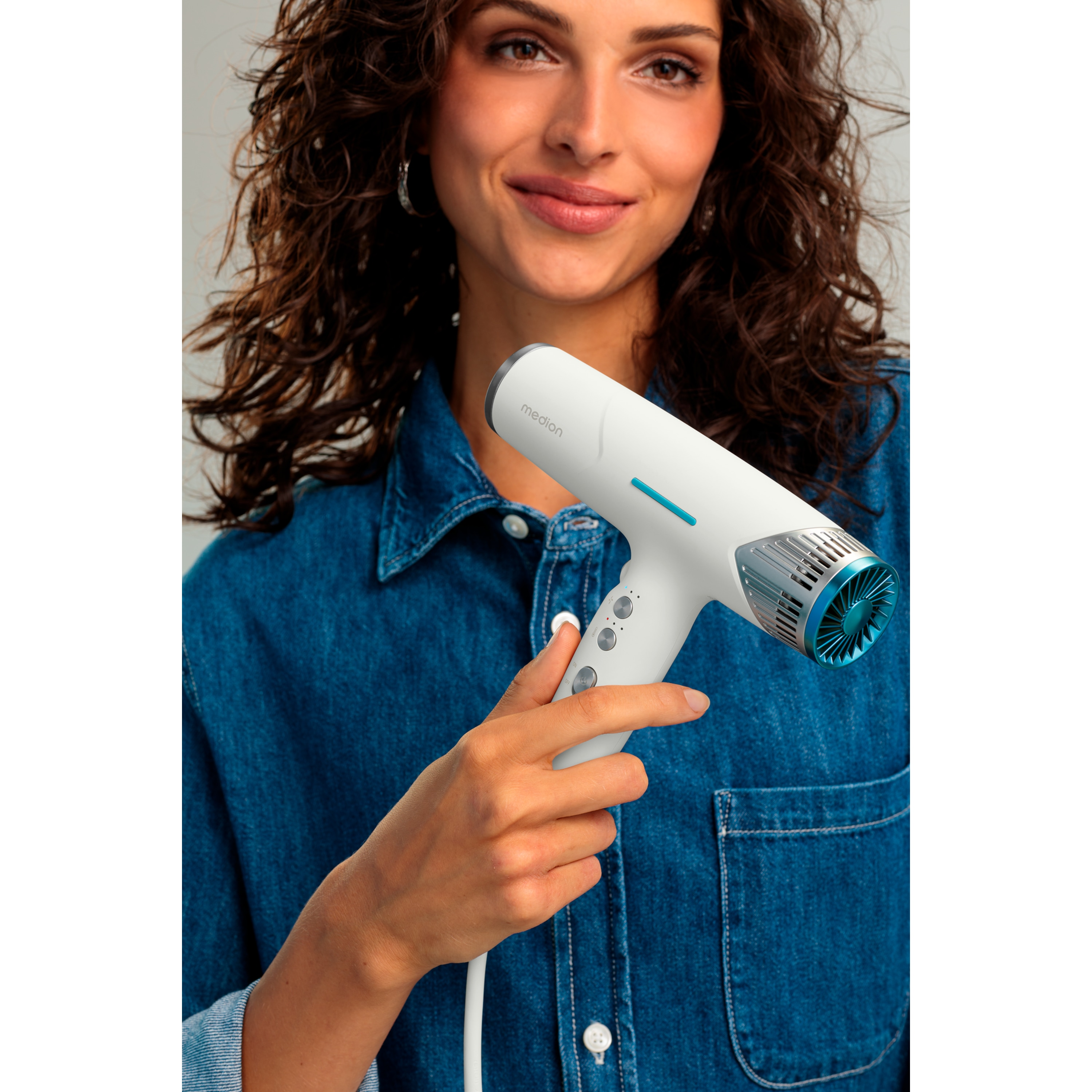 Kobieta trzyma białą suszarkę. Ma na sobie niebieską koszulę dżinsową i kręcone włosy. Tło neutralne.