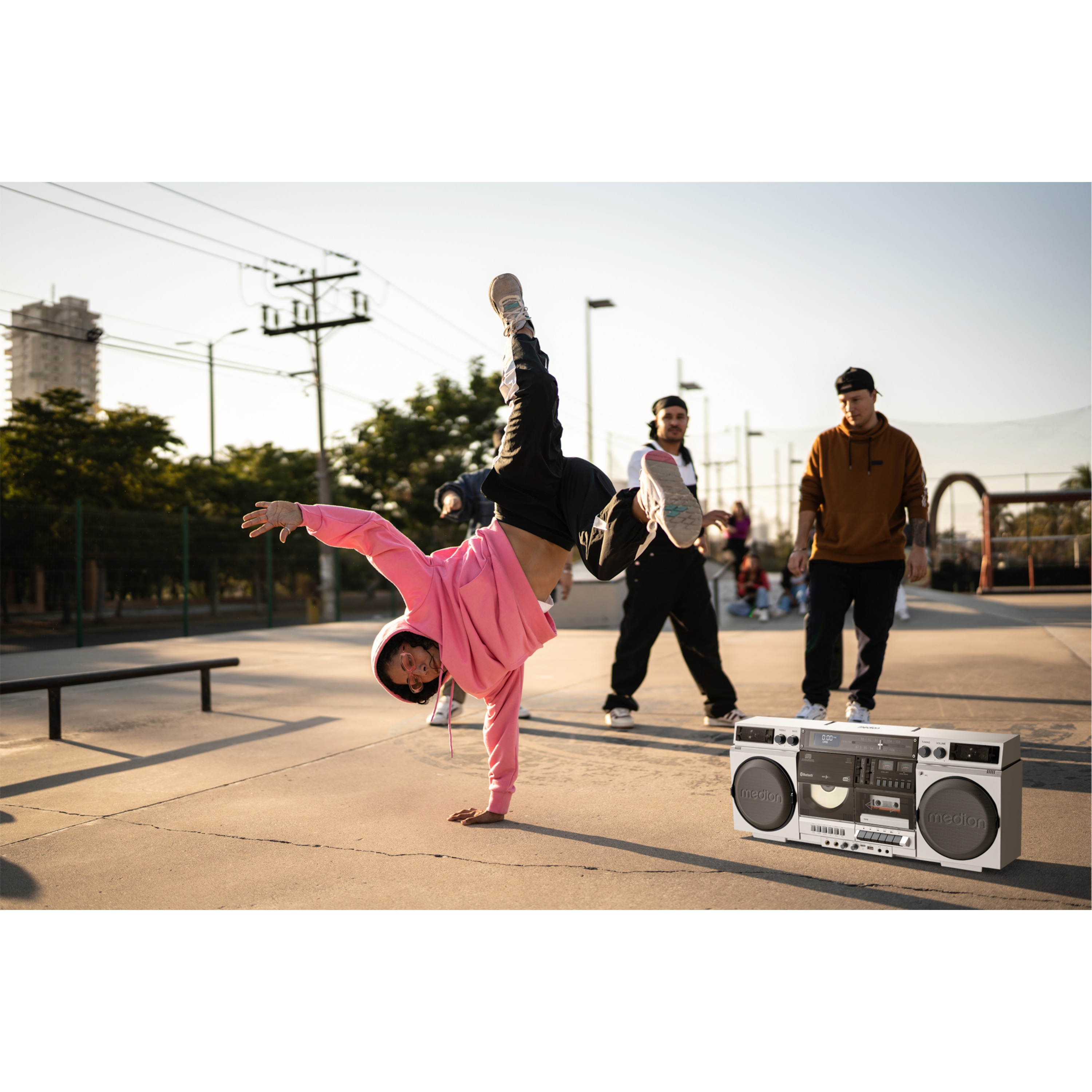 Ein Breakdancer balanciert auf einer Hand. Zwei Männer stehen in der Nähe. Eine Boombox steht im Vordergrund.