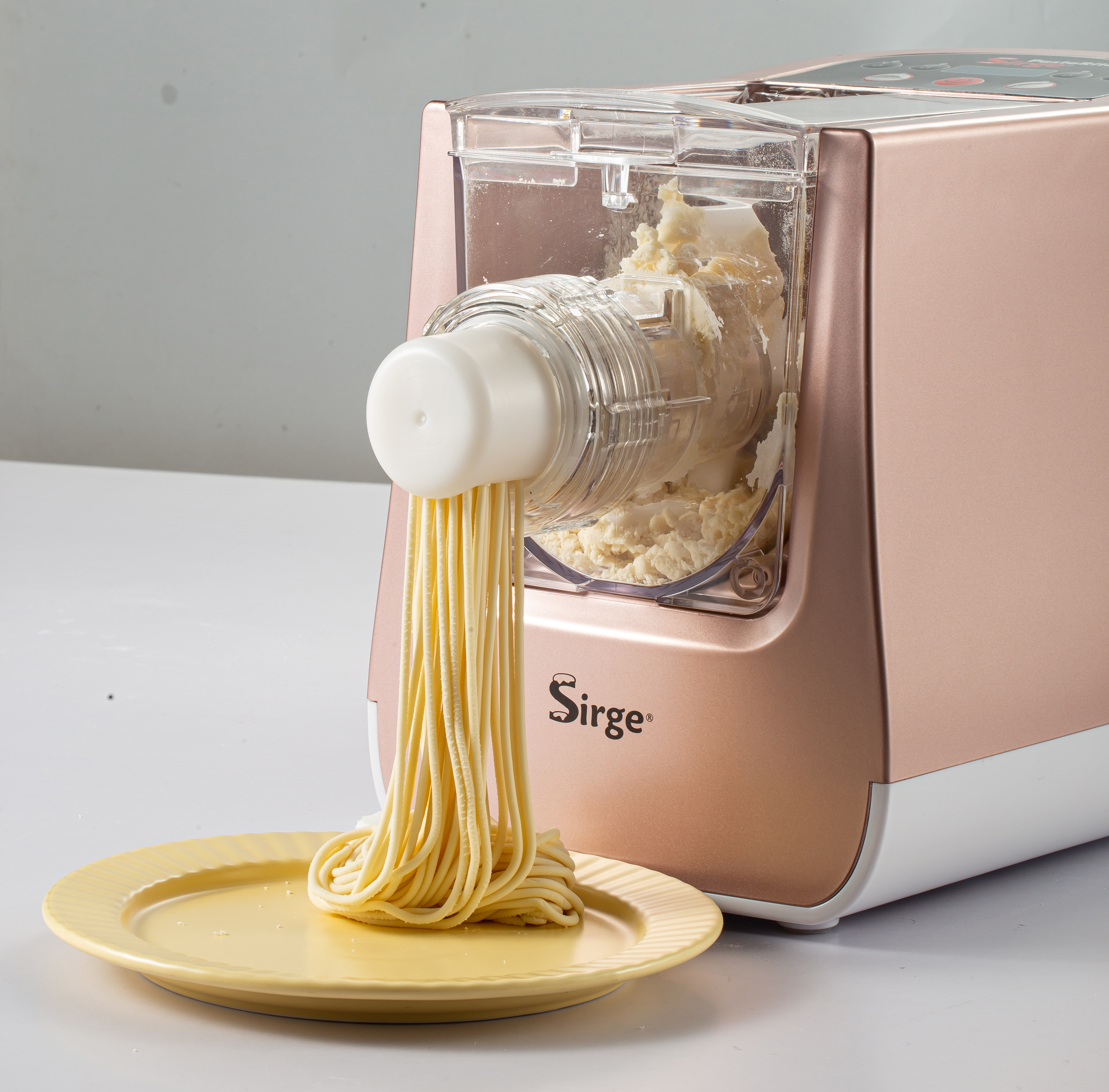 Una macchina per la pasta sta estrudendo pasta fresca su un piatto giallo. La macchina è oro rosa e bianca.
