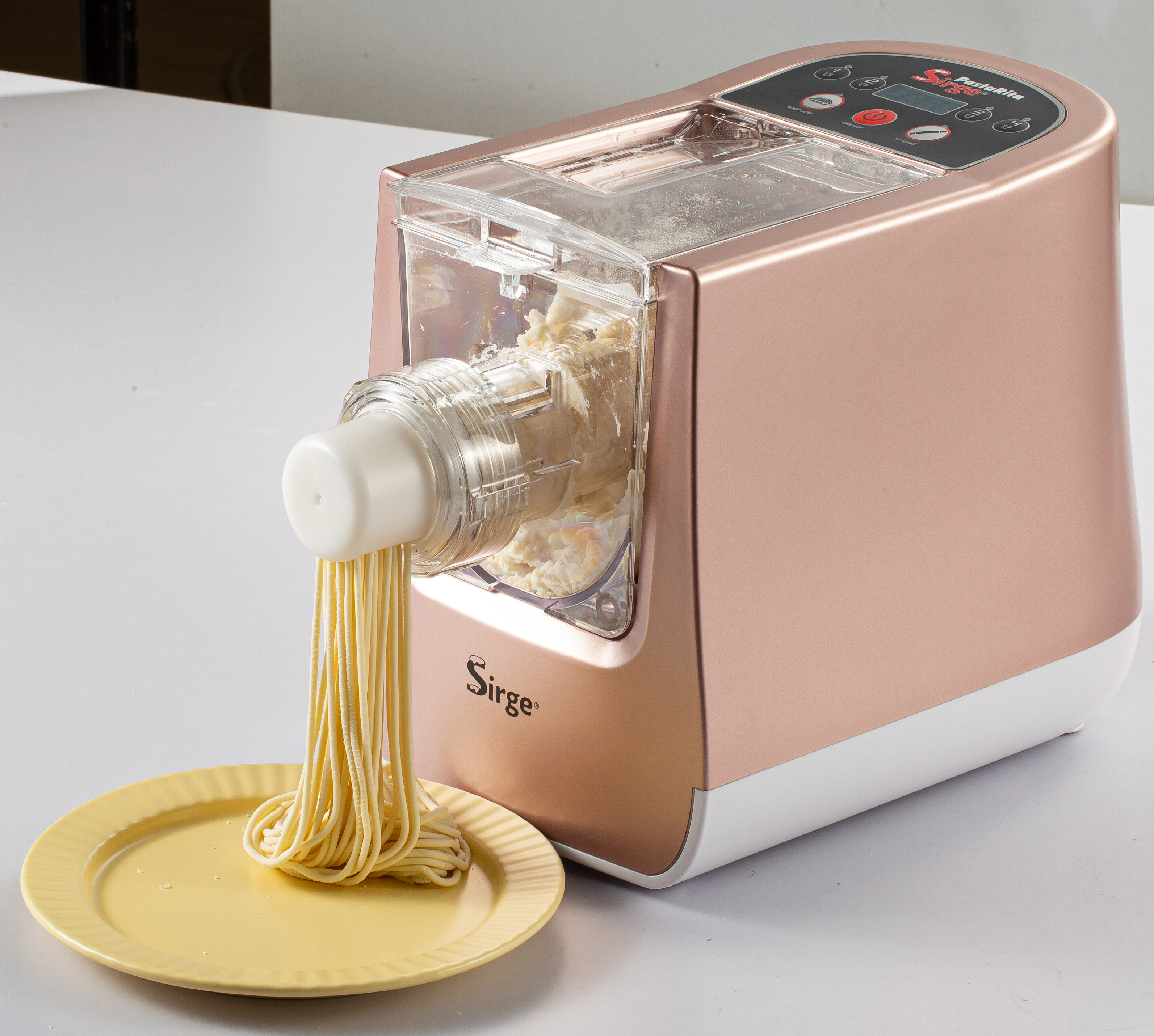 Una macchina per la pasta sta facendo spaghetti. Gli spaghetti cadono su un piatto giallo. La macchina è ramata e bianca.