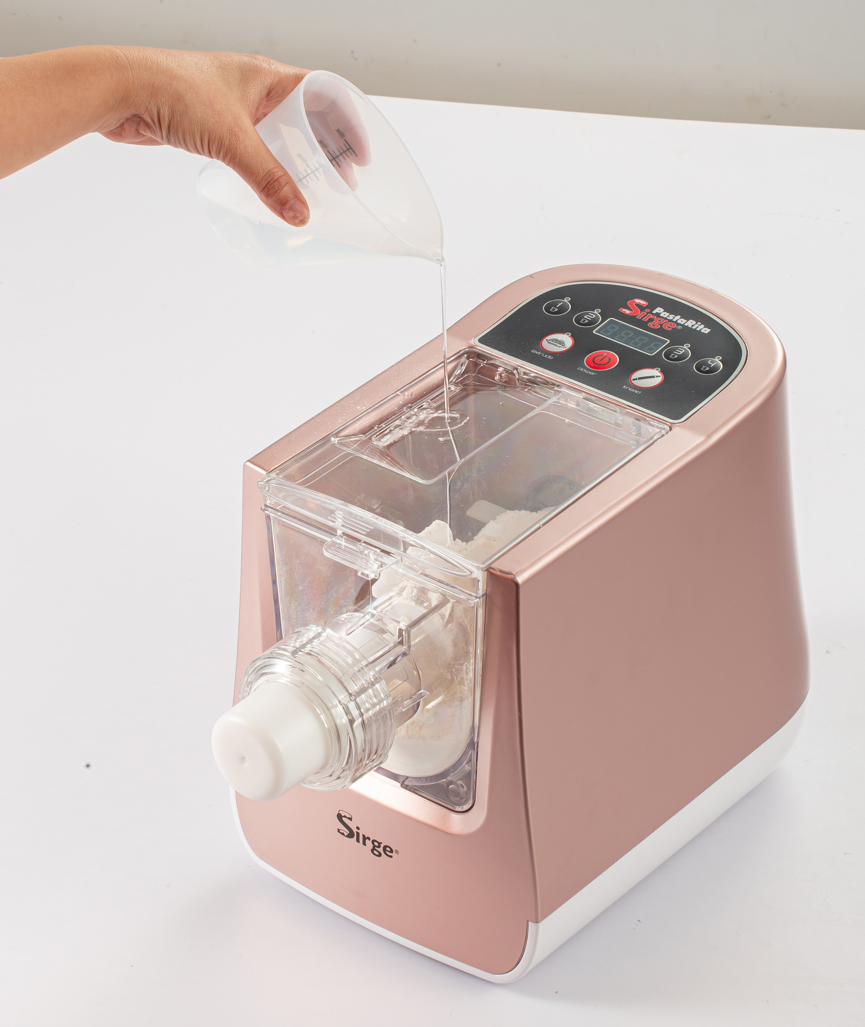 Una persona sta versando acqua in una macchina per la pasta color oro rosa. La macchina è su una superficie bianca.