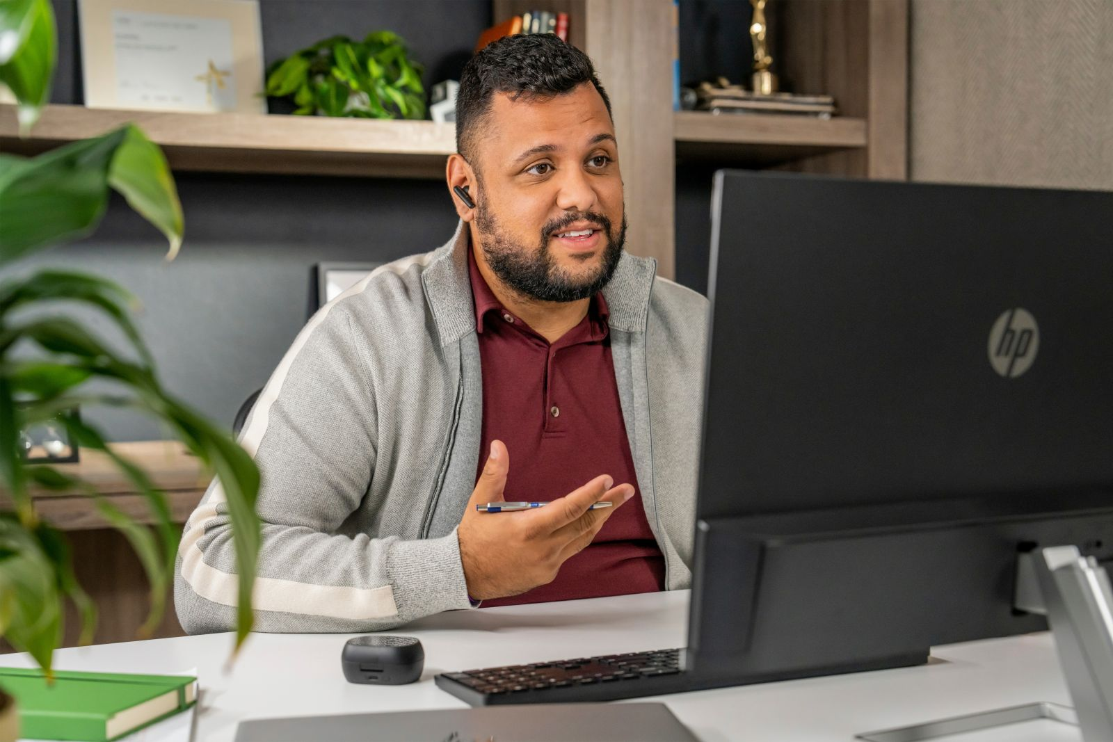 Ein Mann in grauer Jacke und rotem Hemd mit Ohrhörern telefoniert am Schreibtisch mit Monitor, Tastatur und Lautsprecher.