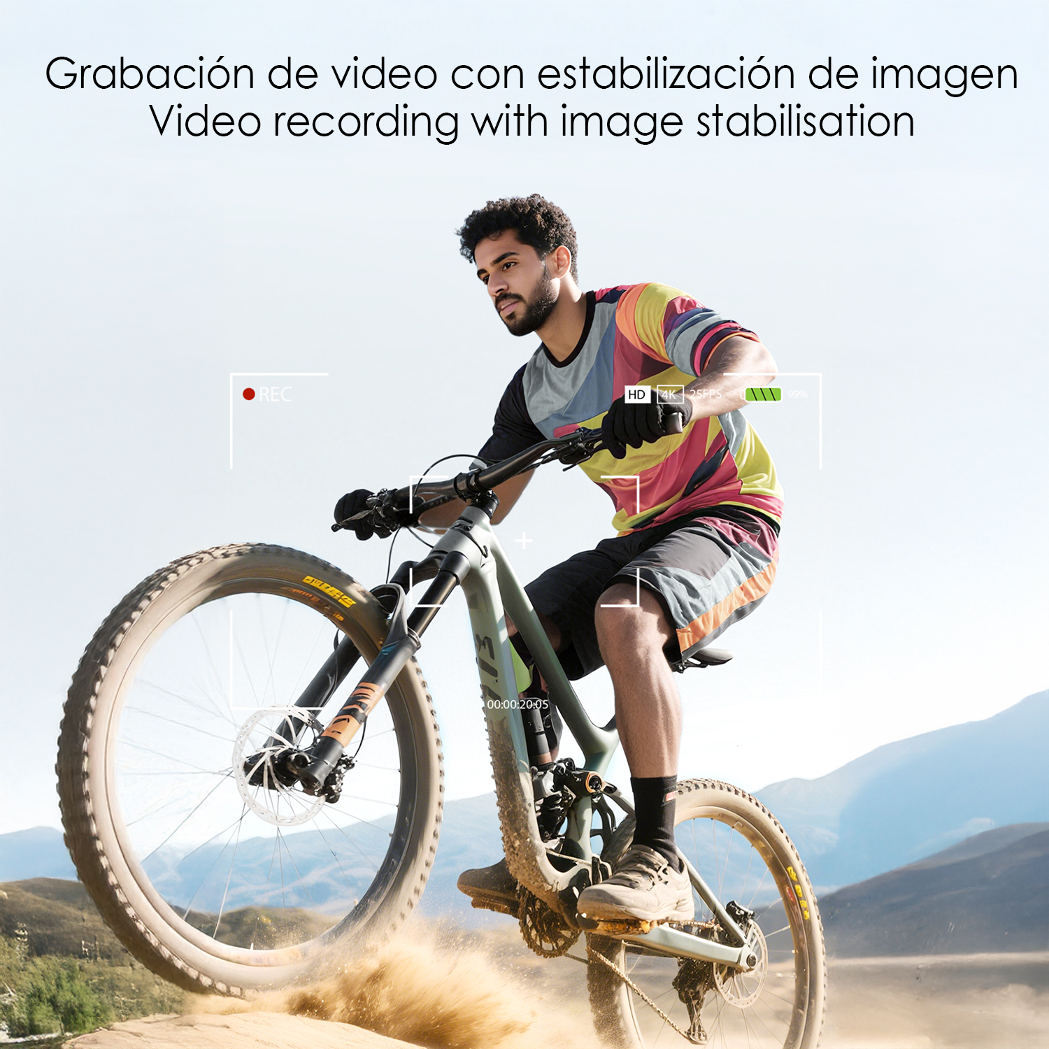 Hombre montando en bicicleta de montaña, levantando polvo. Usa camisa colorida y pantalones cortos. El fondo muestra montañas y cielo.