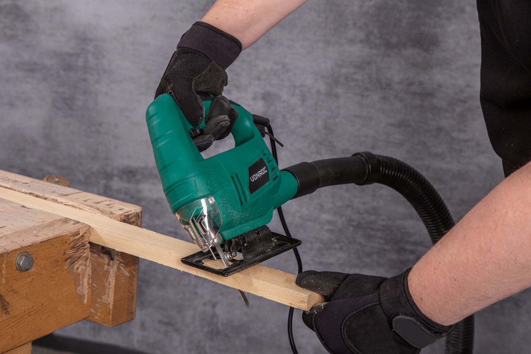 Een persoon gebruikt een groene decoupeerzaag om een houten plank te zagen. Ze dragen zwarte handschoenen. De achtergrond is grijs.