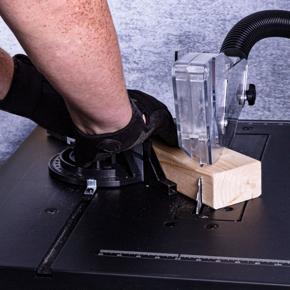 A person wearing a black glove is using a saw to cut a wooden block. The saw has a clear safety shield.