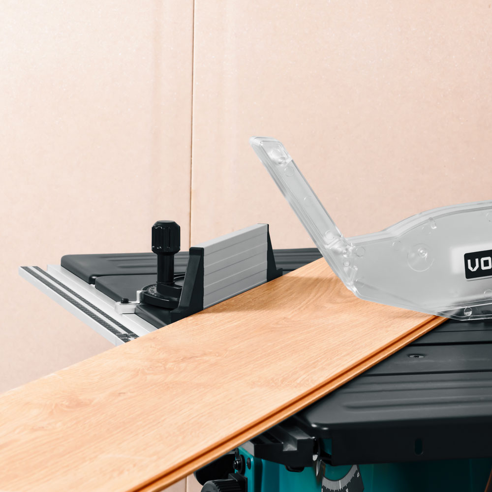A close-up of a table saw with a wooden plank being cut. The saw has a clear safety guard. The background is a neutral wall.