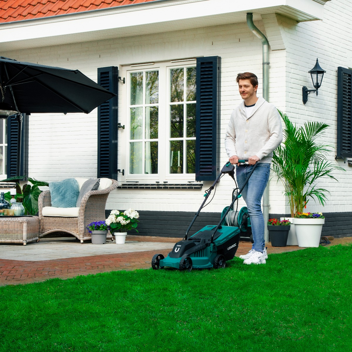 Een man maait een groen gazon voor een wit bakstenen huis met zwarte luiken. Een grasmaaier en planten zijn zichtbaar.