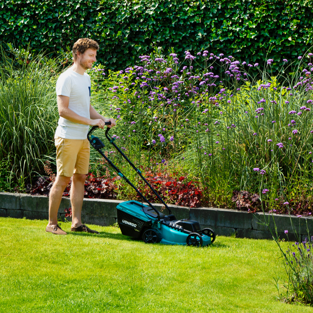 Een man maait een groen gazon met een turquoise grasmaaier. Paarse bloemen en groene struiken op de achtergrond. De man draagt een korte broek en een wit overhemd.